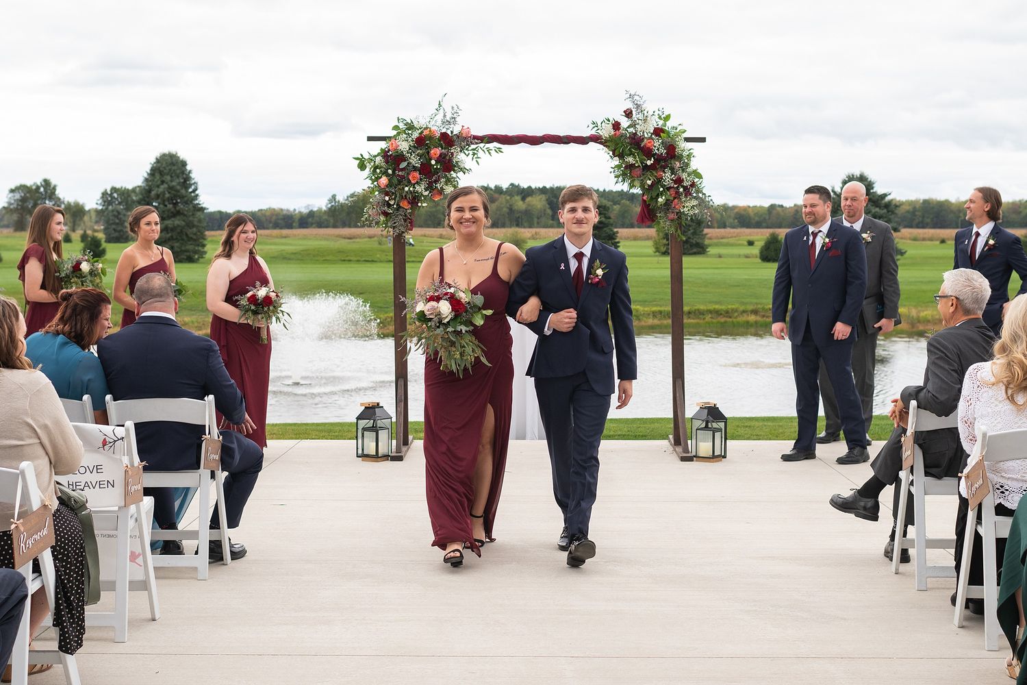 Bridesmaid in burgundy gown escorts groomsman down aisle after outdoor wedding ceremony near scenic pond.