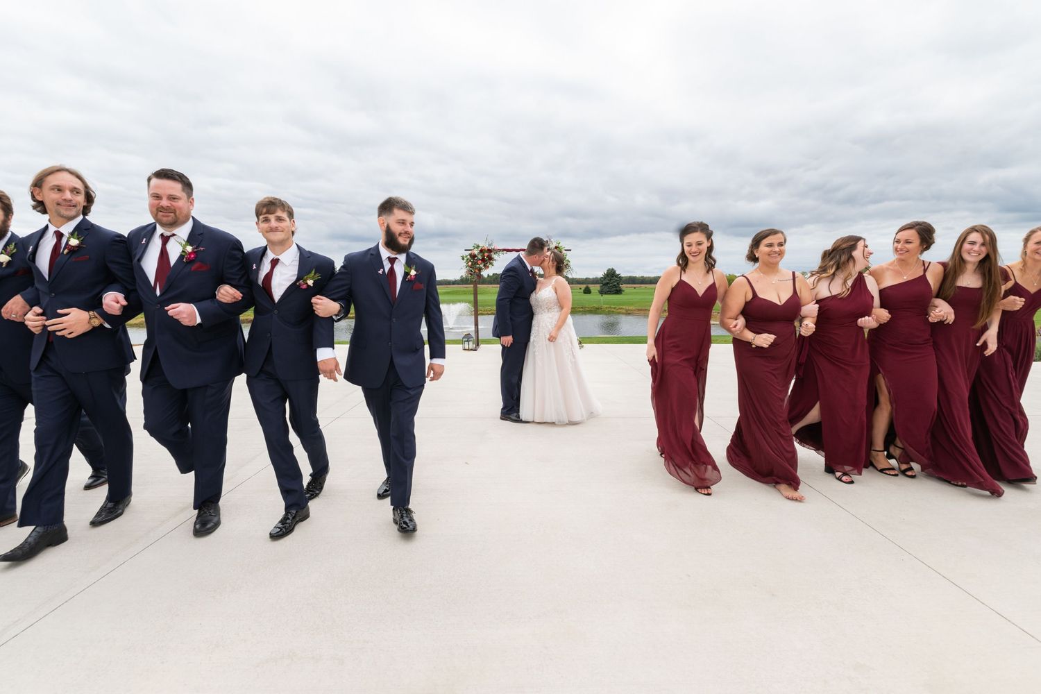 Wedding party in navy suits and burgundy gowns walking outdoors with bride and groom centered in background.