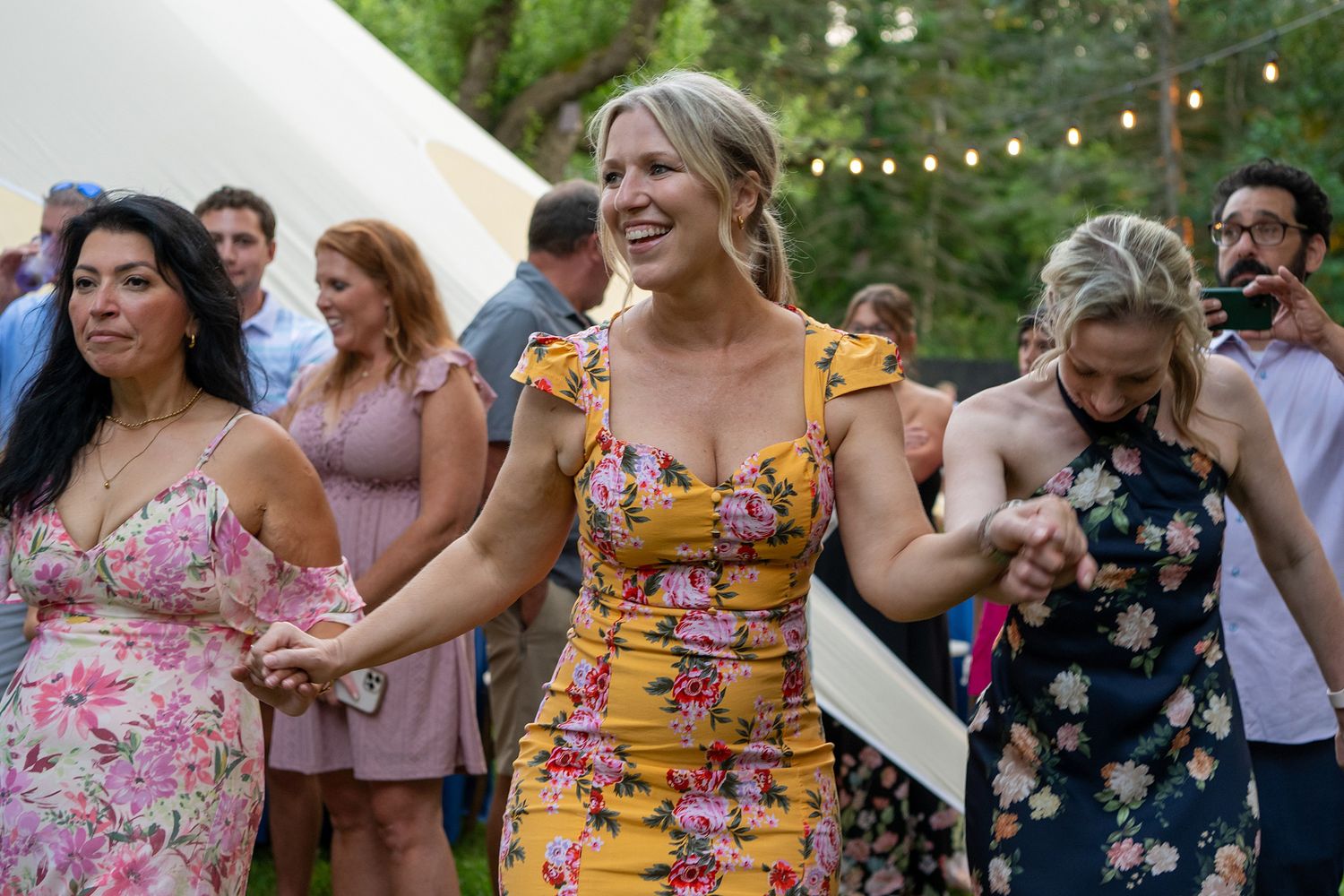 Wedding guests in floral dresses dance together at an outdoor reception with string lights overhead.