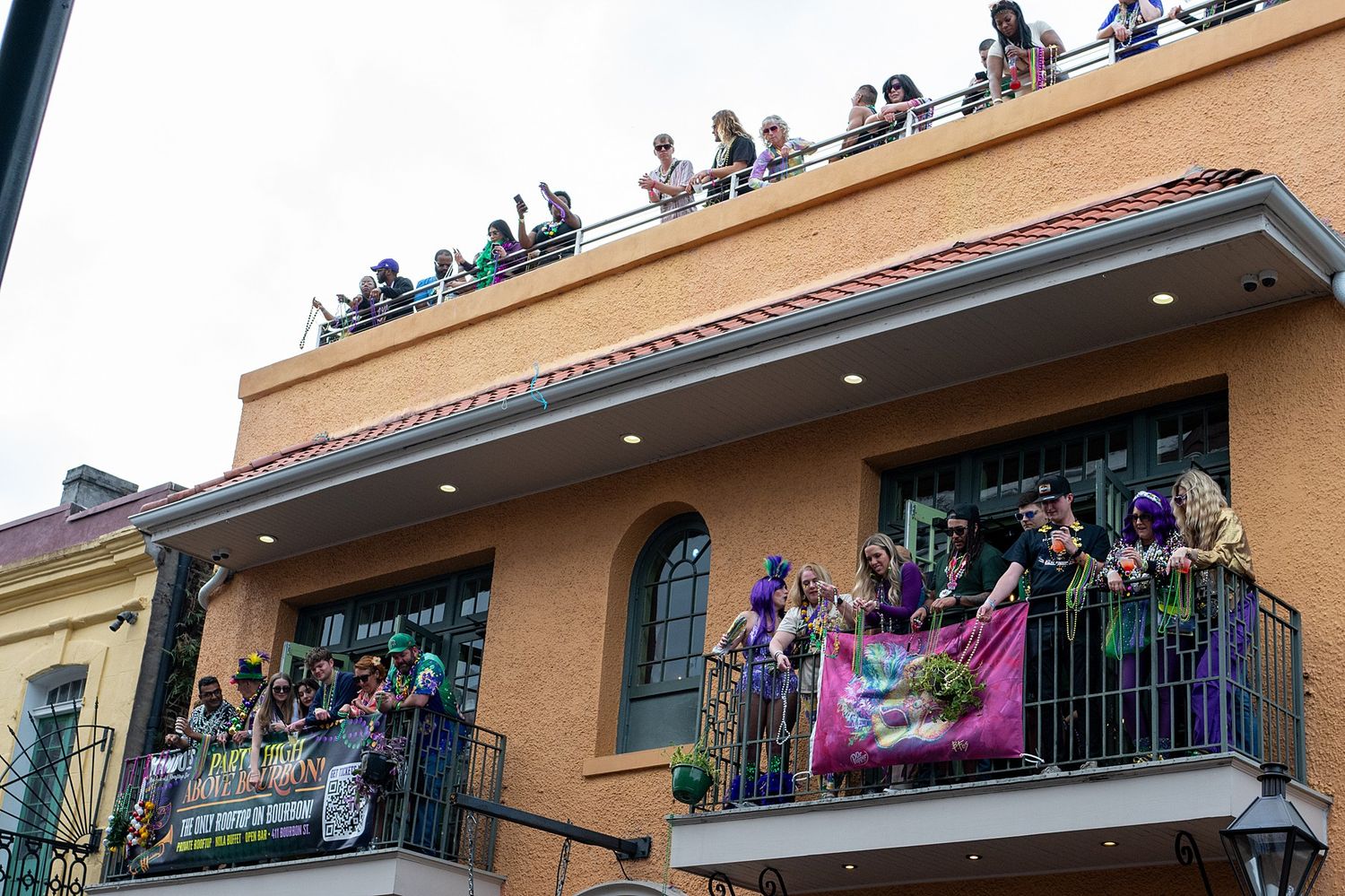 Crowded balcony overlooking Bourbon Street filled with Mardi Gras party spectators.