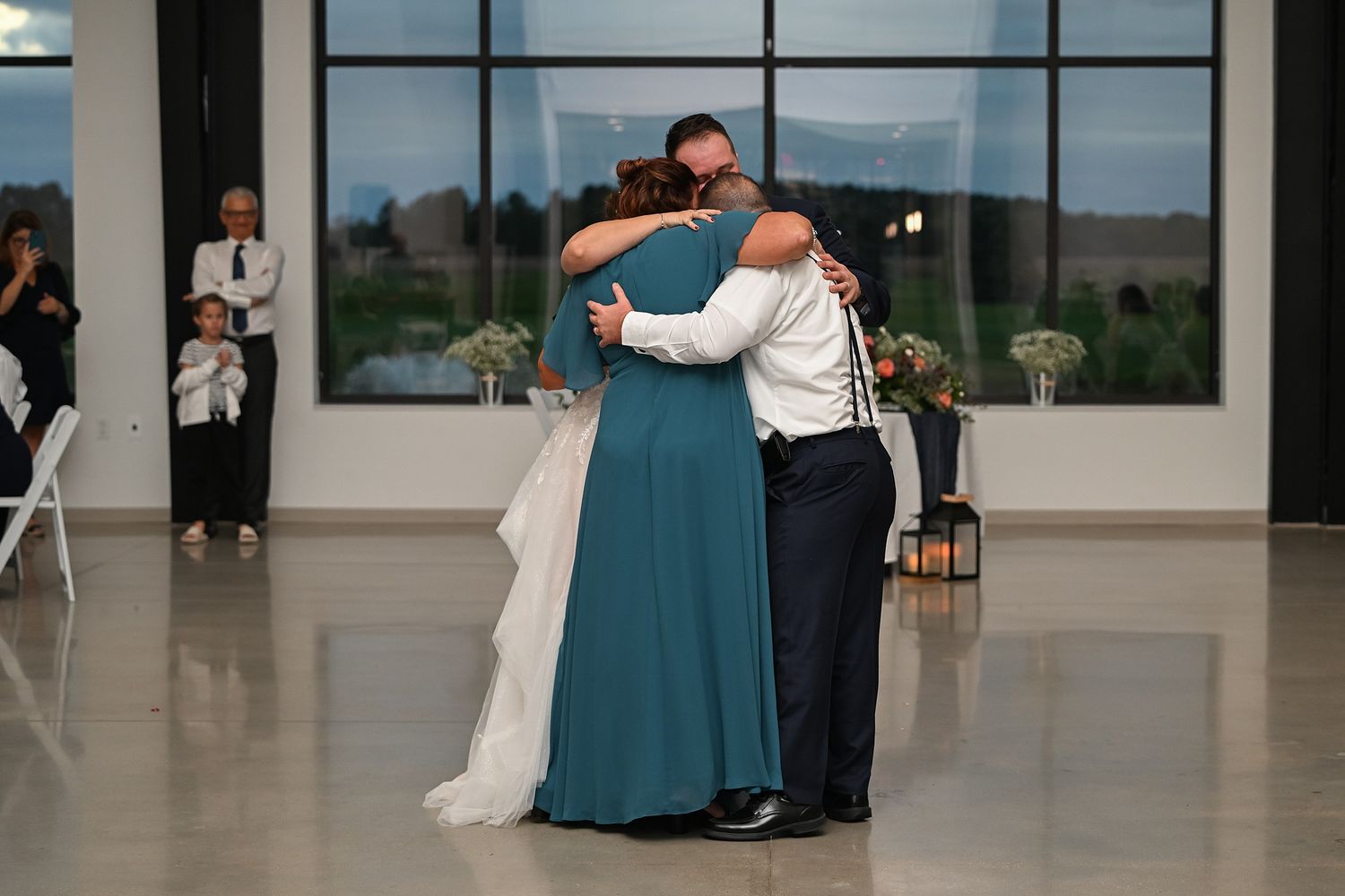 Bride and groom share a heartfelt three-way hug with a guest in teal dress on dance floor.