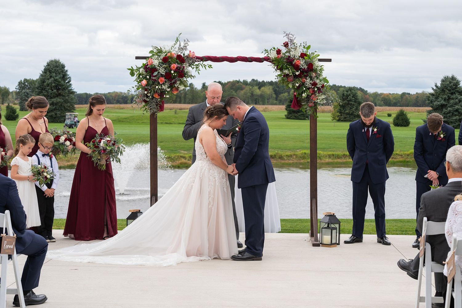 Bride and groom share their first kiss under a floral arch at an outdoor wedding ceremony beside a serene pond.