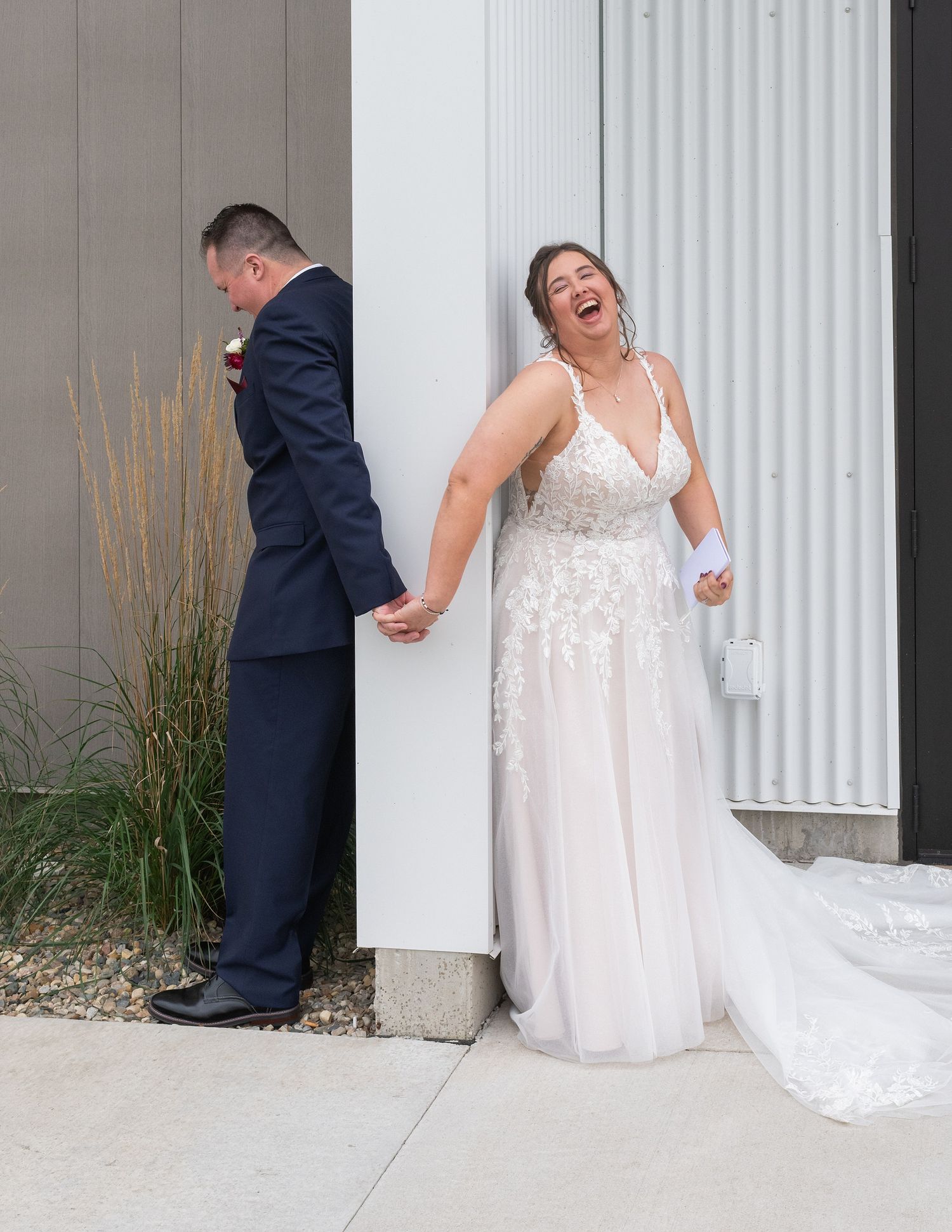 Bride laughing joyfully in white lace gown holds groom's hand around a corner in a wedding first look.