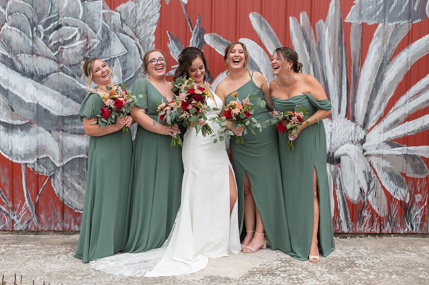 Bride in white gown laughing with three bridesmaids in sage green dresses holding red bouquets against a floral mural.