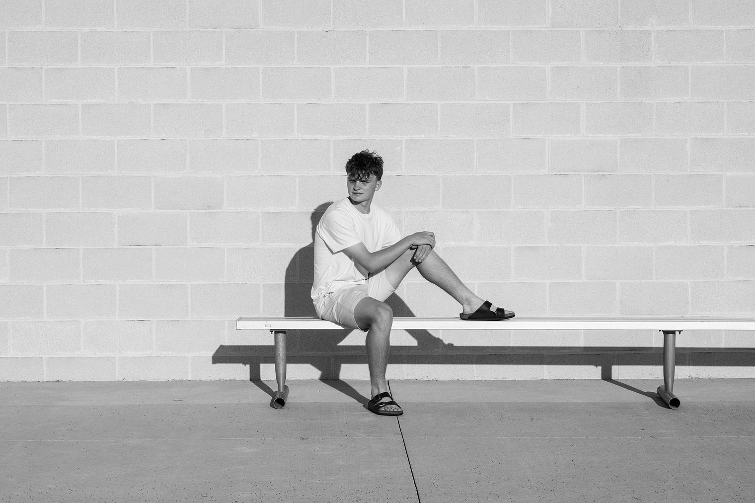 A person in casual attire sits on a white bench against a light colored wall.