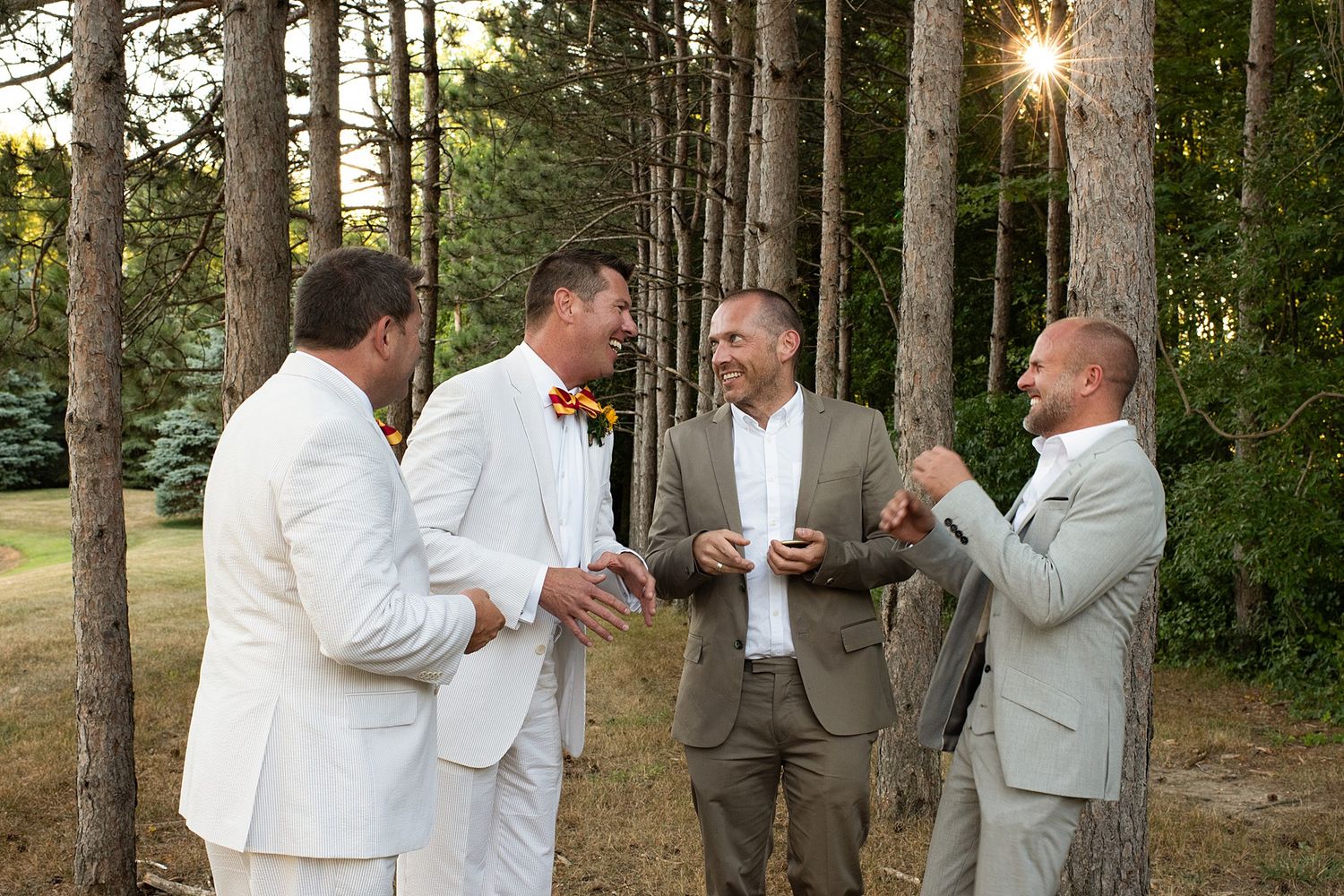 Four men in wedding attire laughing and celebrating outdoors among tall pine trees on a sunny day.
