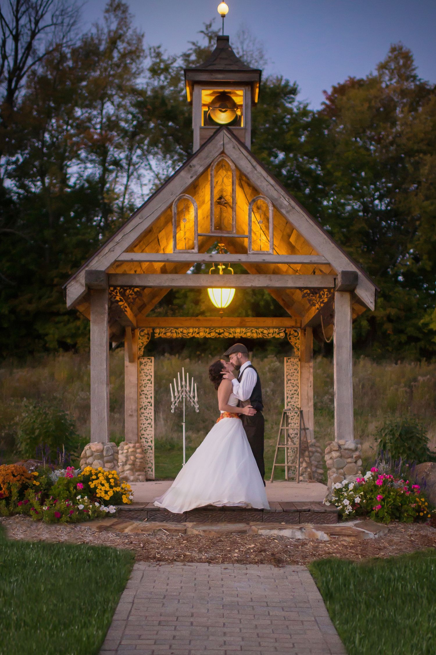 Bride and groom share a romantic kiss under a glowing wooden chapel gazebo surrounded by autumn flowers at dusk.