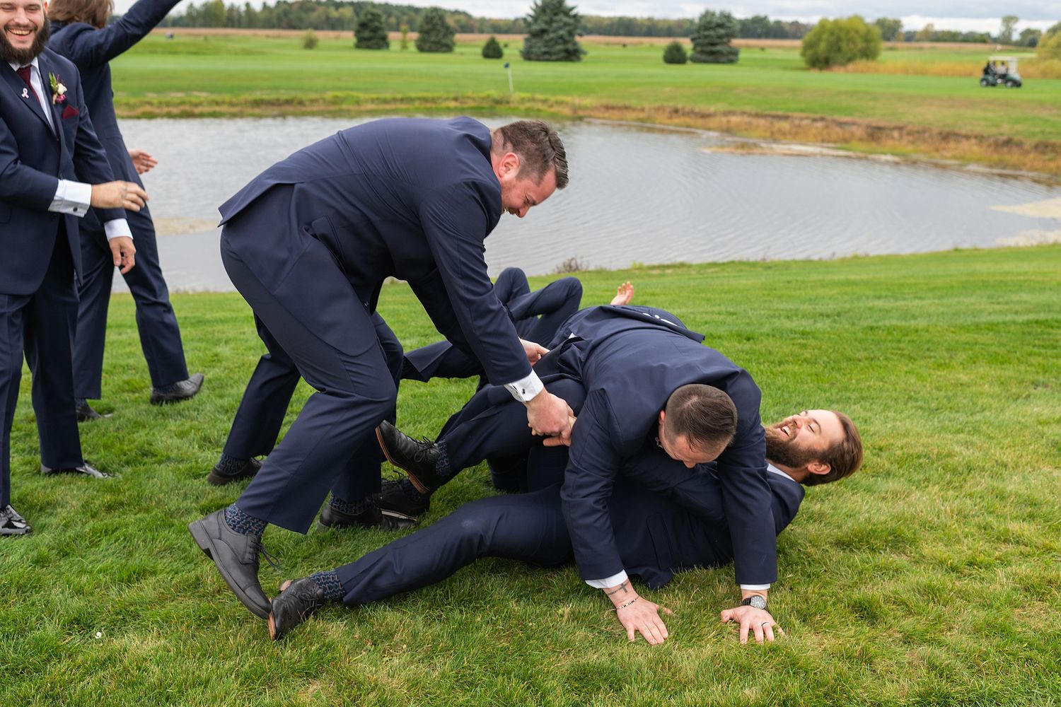 Men in navy suits playfully wrestling on green grass near a pond at an outdoor wedding celebration.