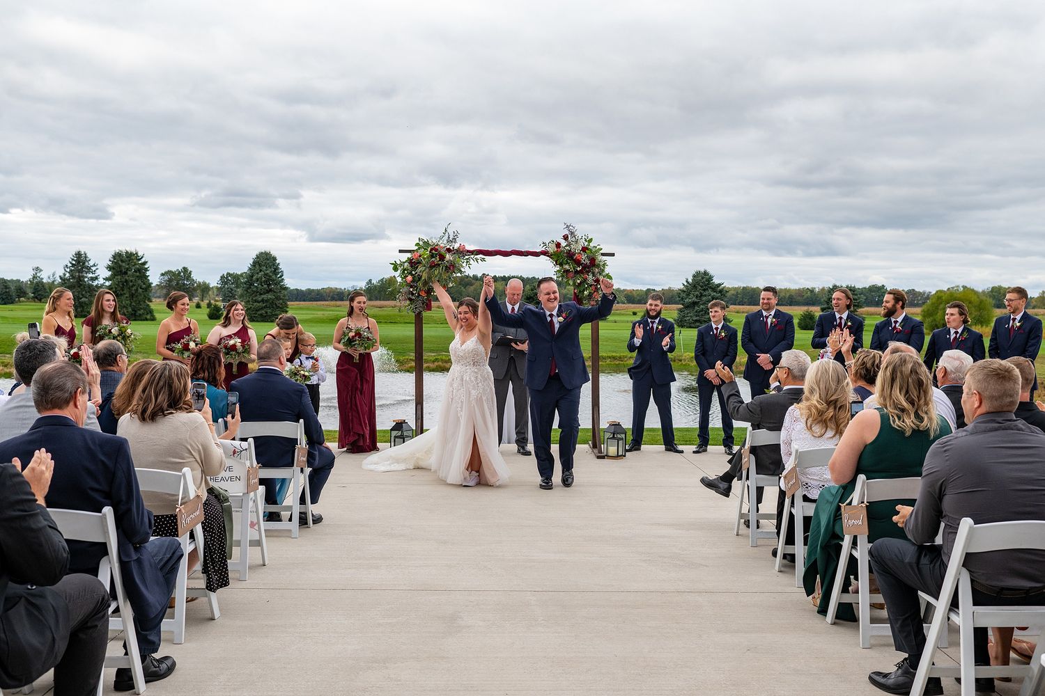 Newlyweds walk down the aisle at outdoor wedding ceremony with floral arch backdrop.