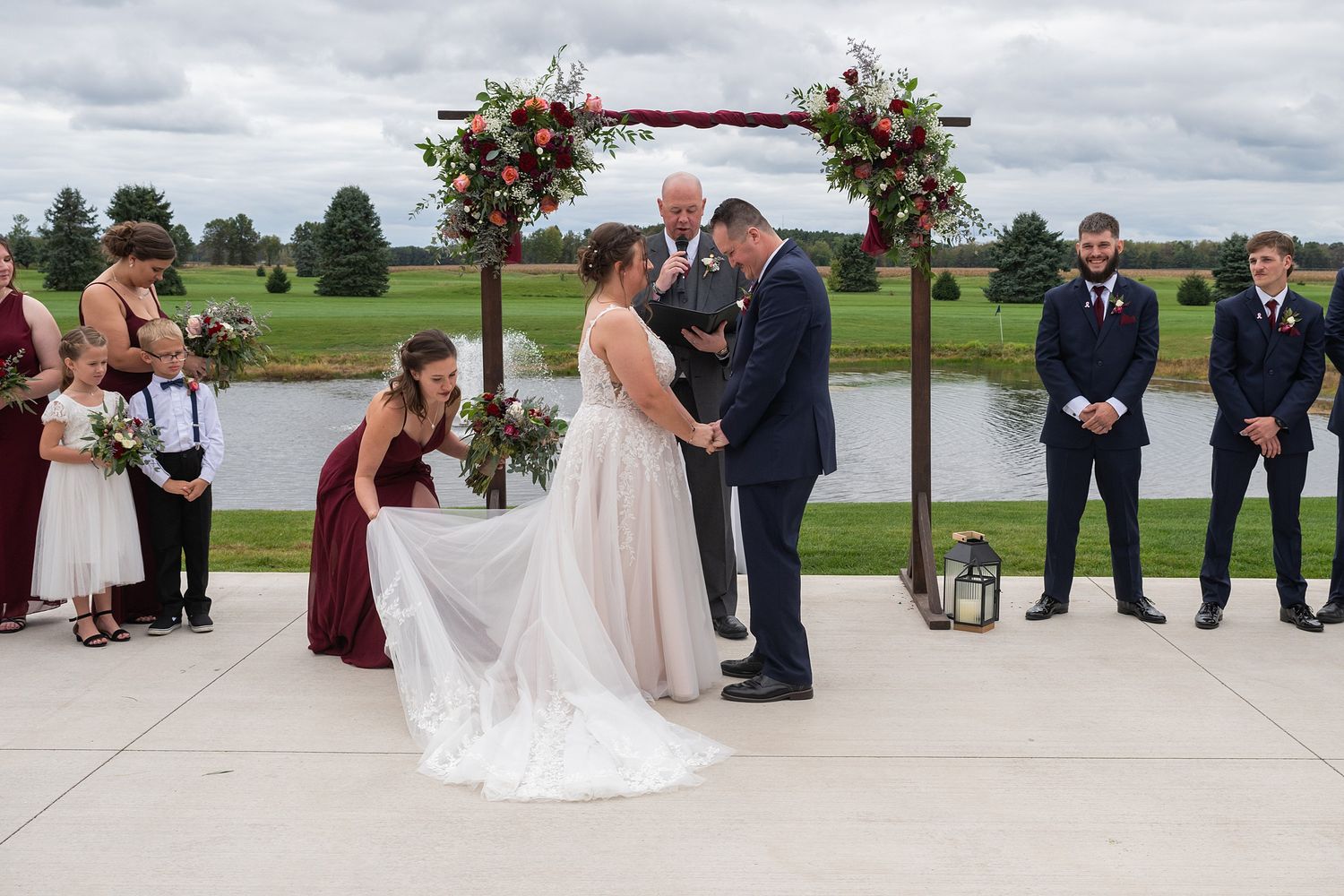 Outdoor wedding ceremony at pond venue with bride, groom, officiant, and wedding party in burgundy.