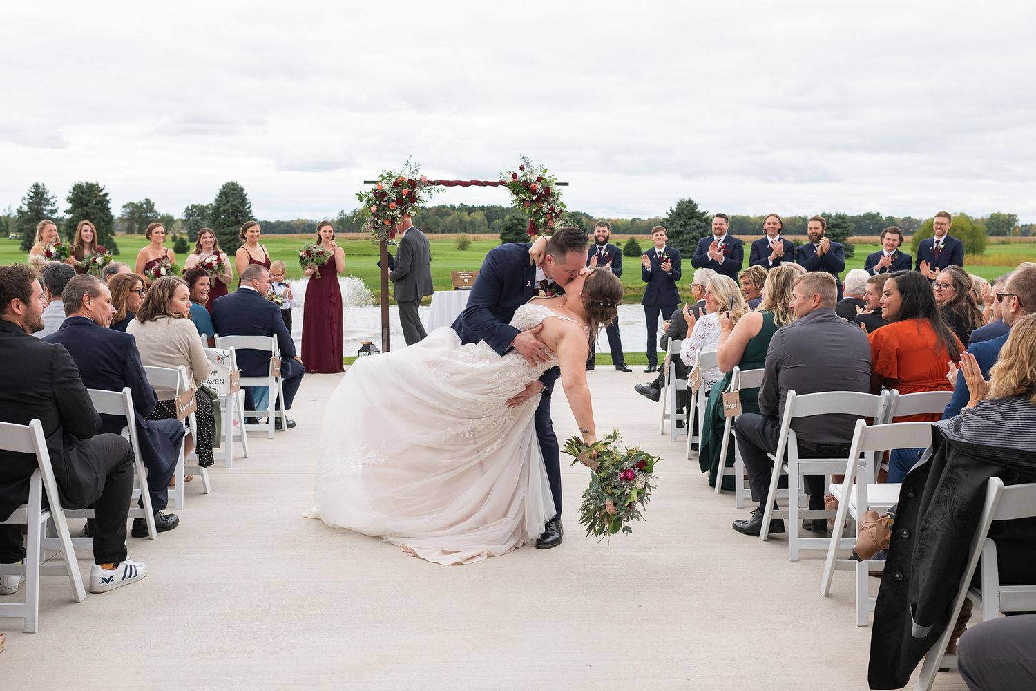 Groom dips bride for romantic kiss in wedding aisle surrounded by cheering guests outdoors.