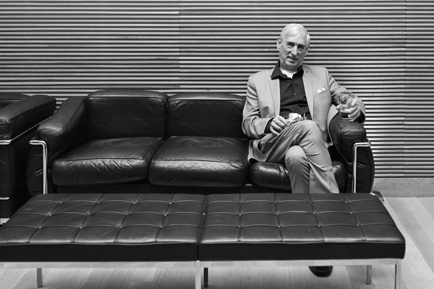 Black and white photo of an elderly man in a suit sitting on a leather sofa in a modern minimalist interior.