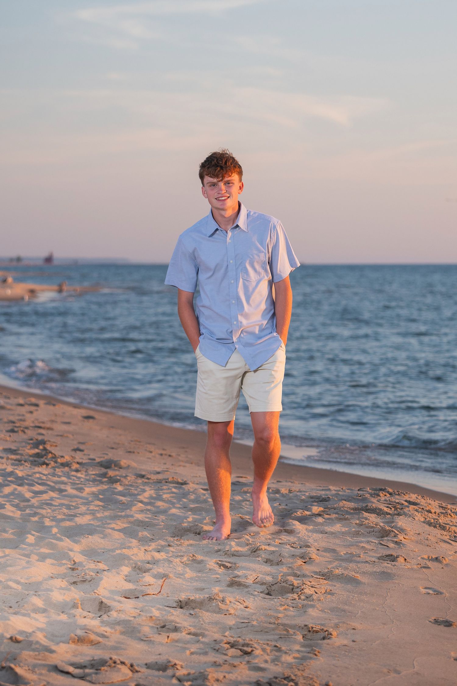 Person in light blue shirt and khaki shorts walking along beach at sunset.