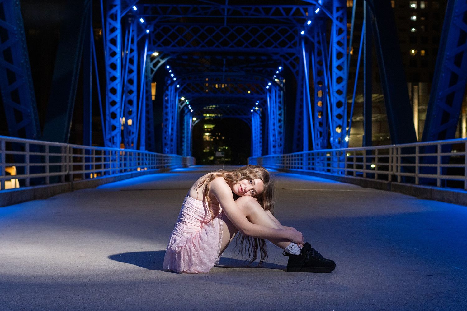 A person in a pink dress sits on a blue-lit bridge walkway at night.