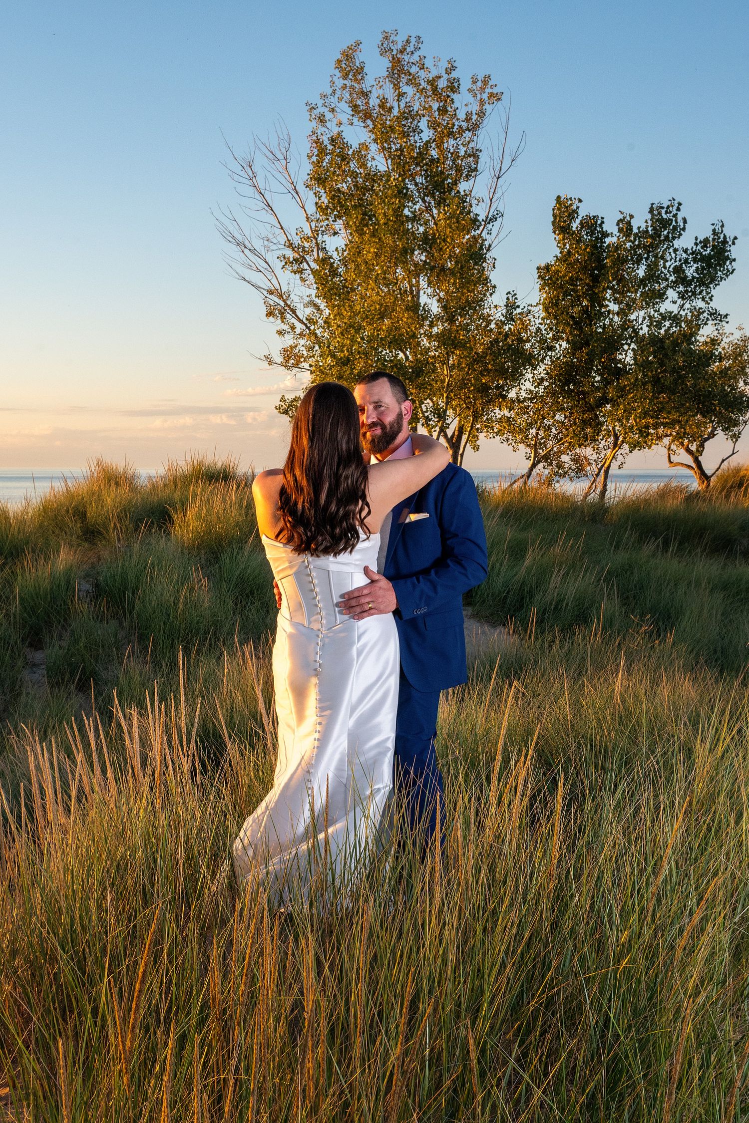 Bride in a white gown and groom in a blue suit embrace in tall beach grass at sunset near a lake, with trees in the background.