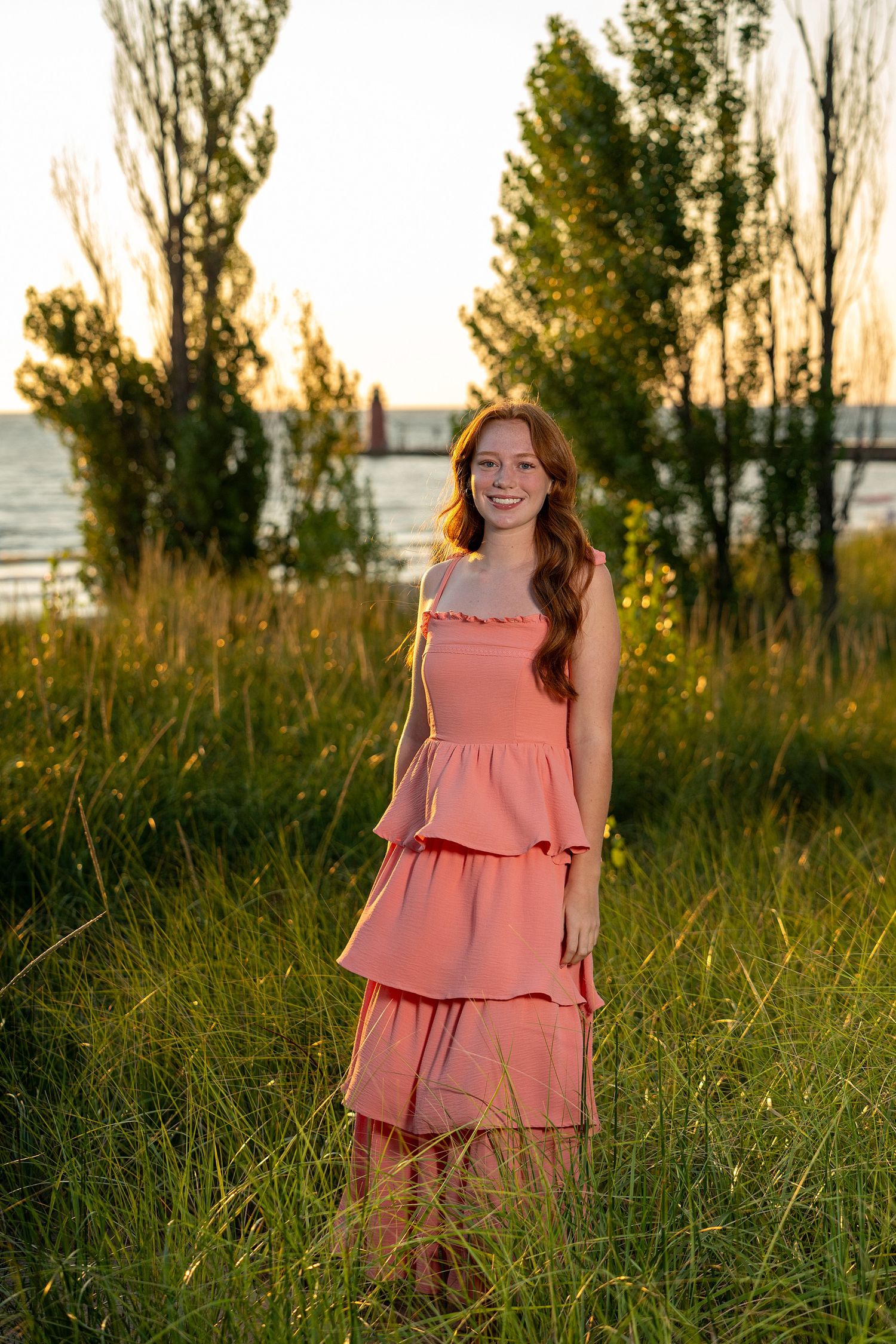 Young woman in pink tiered dress standing in tall grass at golden hour near waterfront.