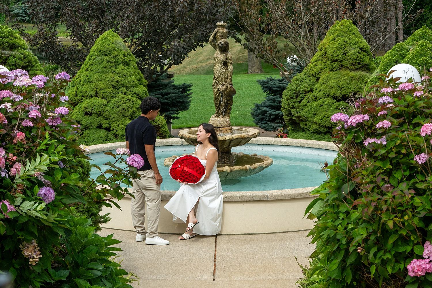 Couple poses by ornate garden fountain surrounded by trimmed hedges and pink hydrangeas, woman holds red heart balloon.