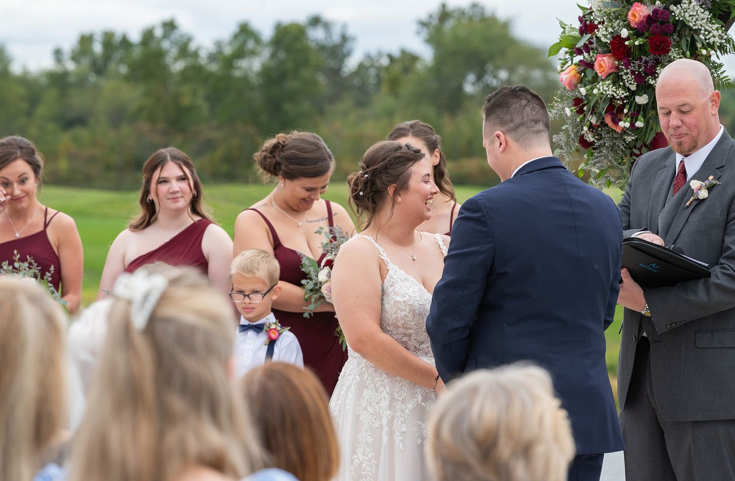 Bride and groom exchange vows while bridesmaids in burgundy dresses look on at outdoor ceremony.