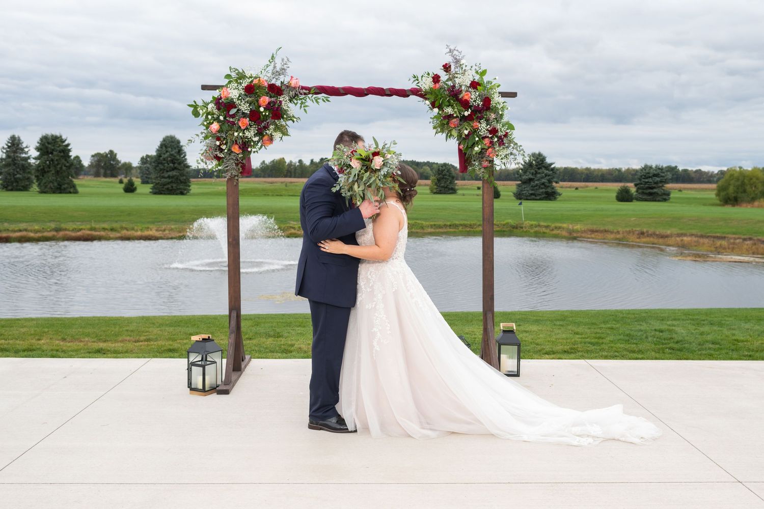 Bride and groom sharing first kiss under a rustic wooden arch adorned with burgundy and blush floral arrangements by a pond.