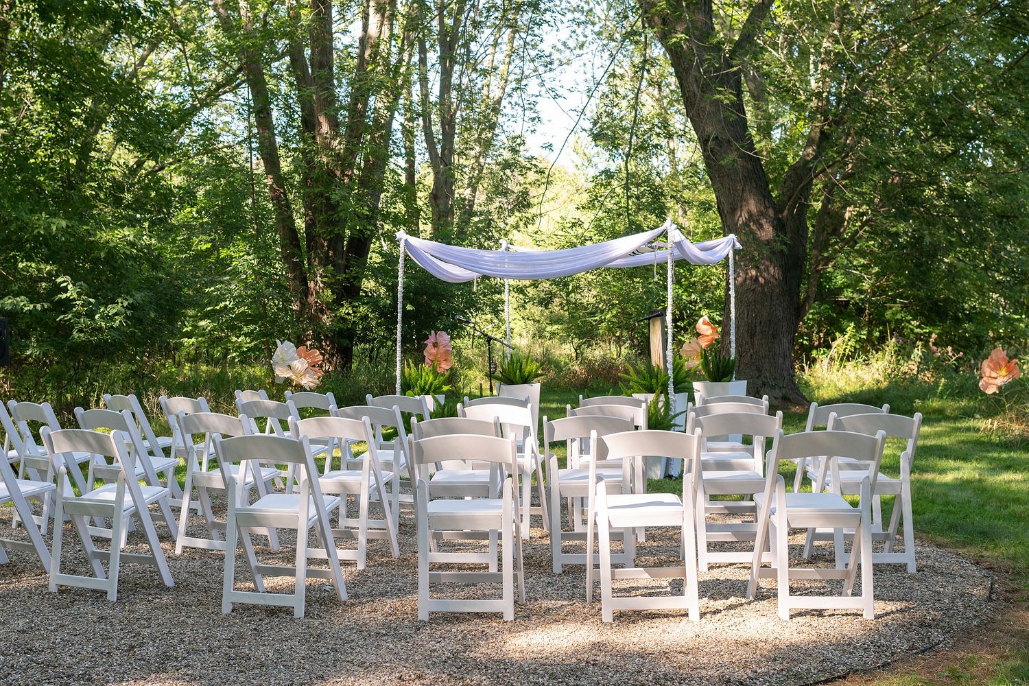 White wooden chairs arranged in rows for an outdoor wedding ceremony under shady trees in a garden setting.