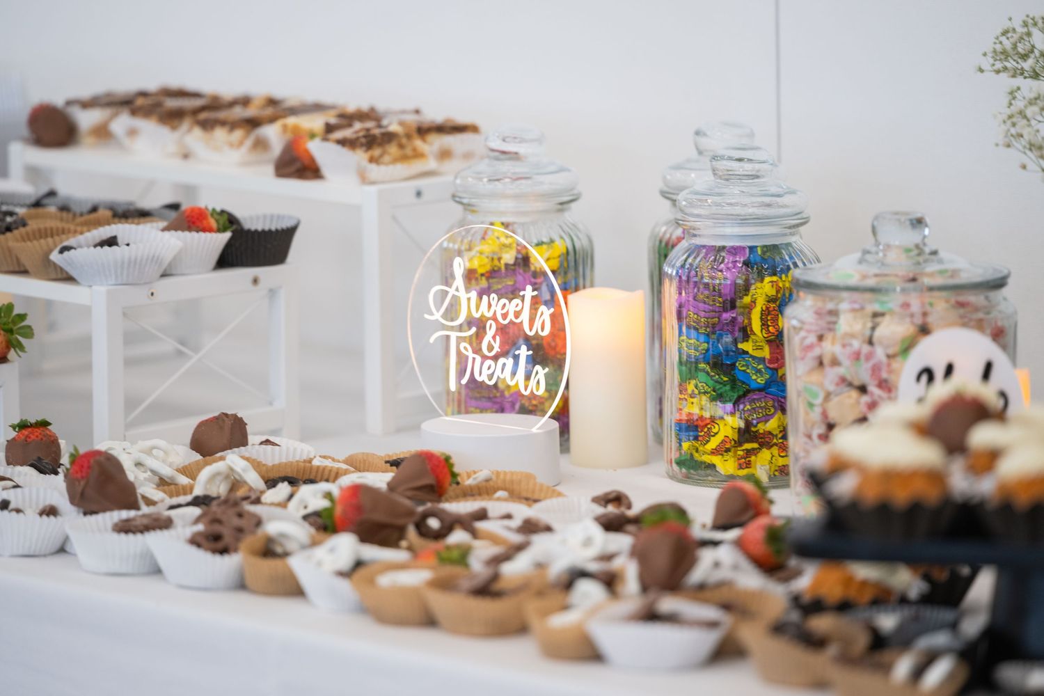 Elegant dessert table with chocolate-dipped strawberries, cupcakes, candy jars, and a Sweet Treats sign at a wedding reception.