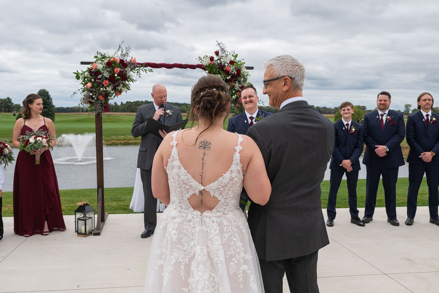 Bride stands at outdoor wedding ceremony altar with groom and wedding party beside a pond.
