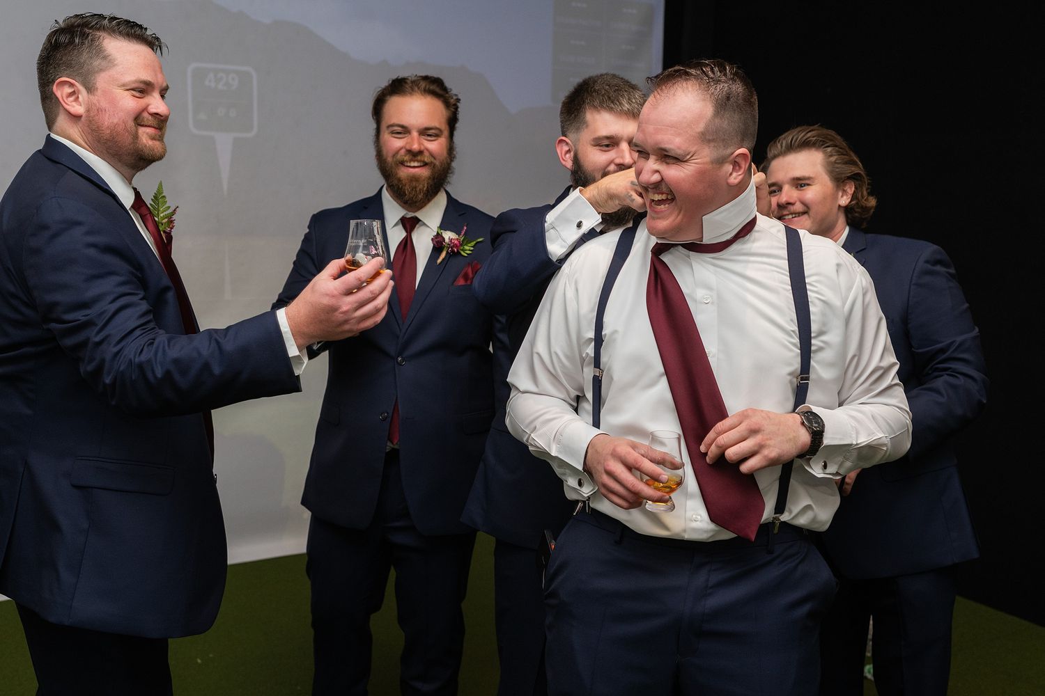 Group of groomsmen in navy suits laughing and joking together at a wedding reception.