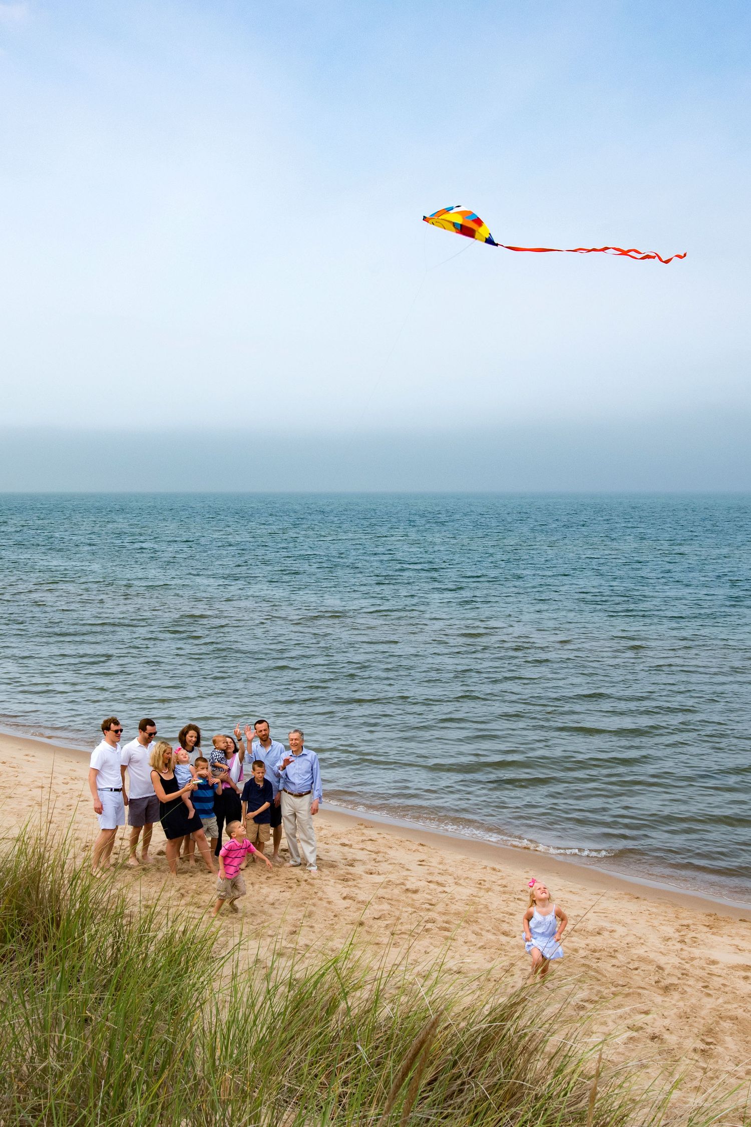 Family group on sandy beach flying a colorful yellow kite with red tail, ocean waves in background, sunny day.