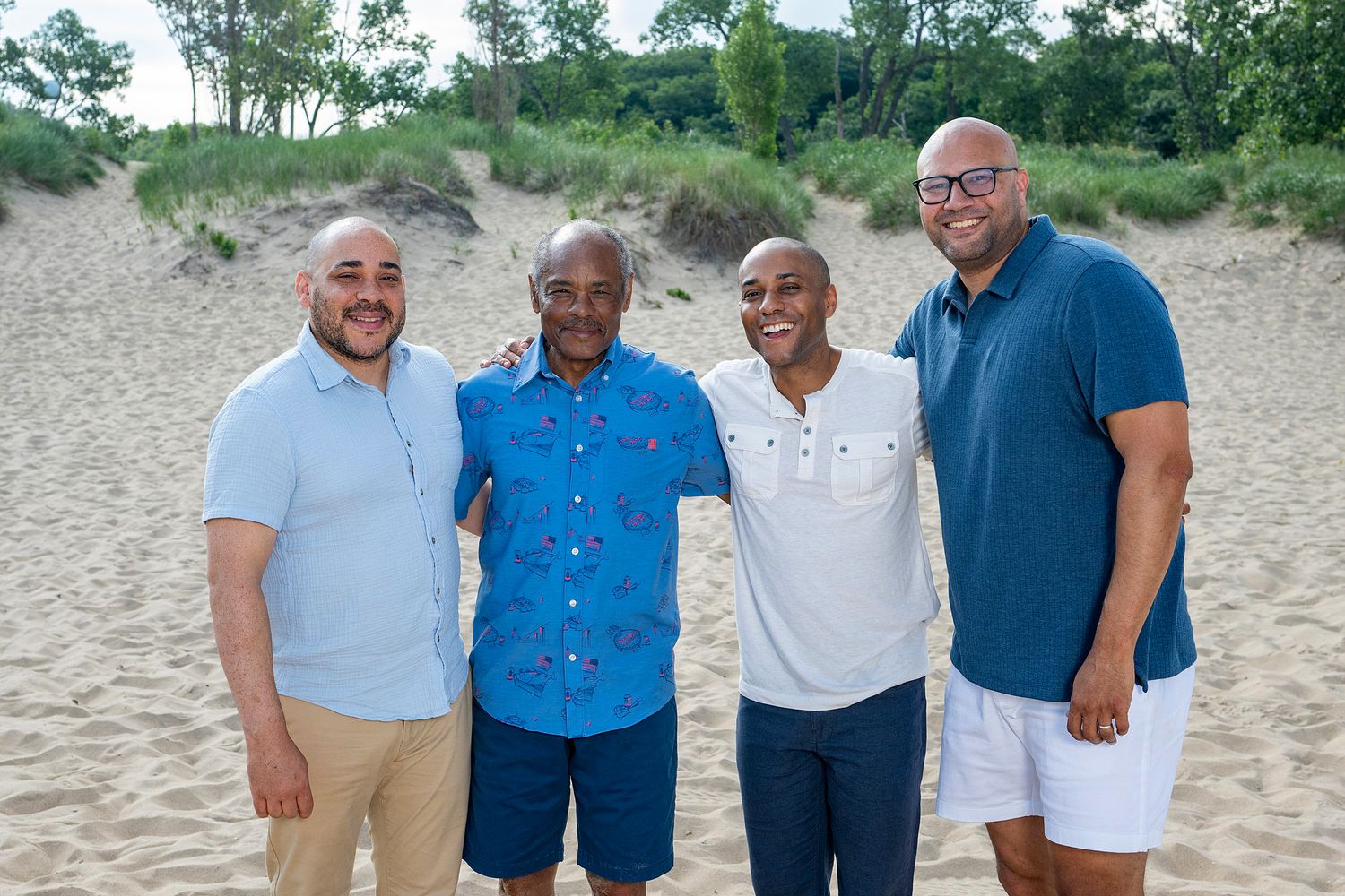 Four smiling men pose together on a sandy beach with green trees and sand dunes in the background.