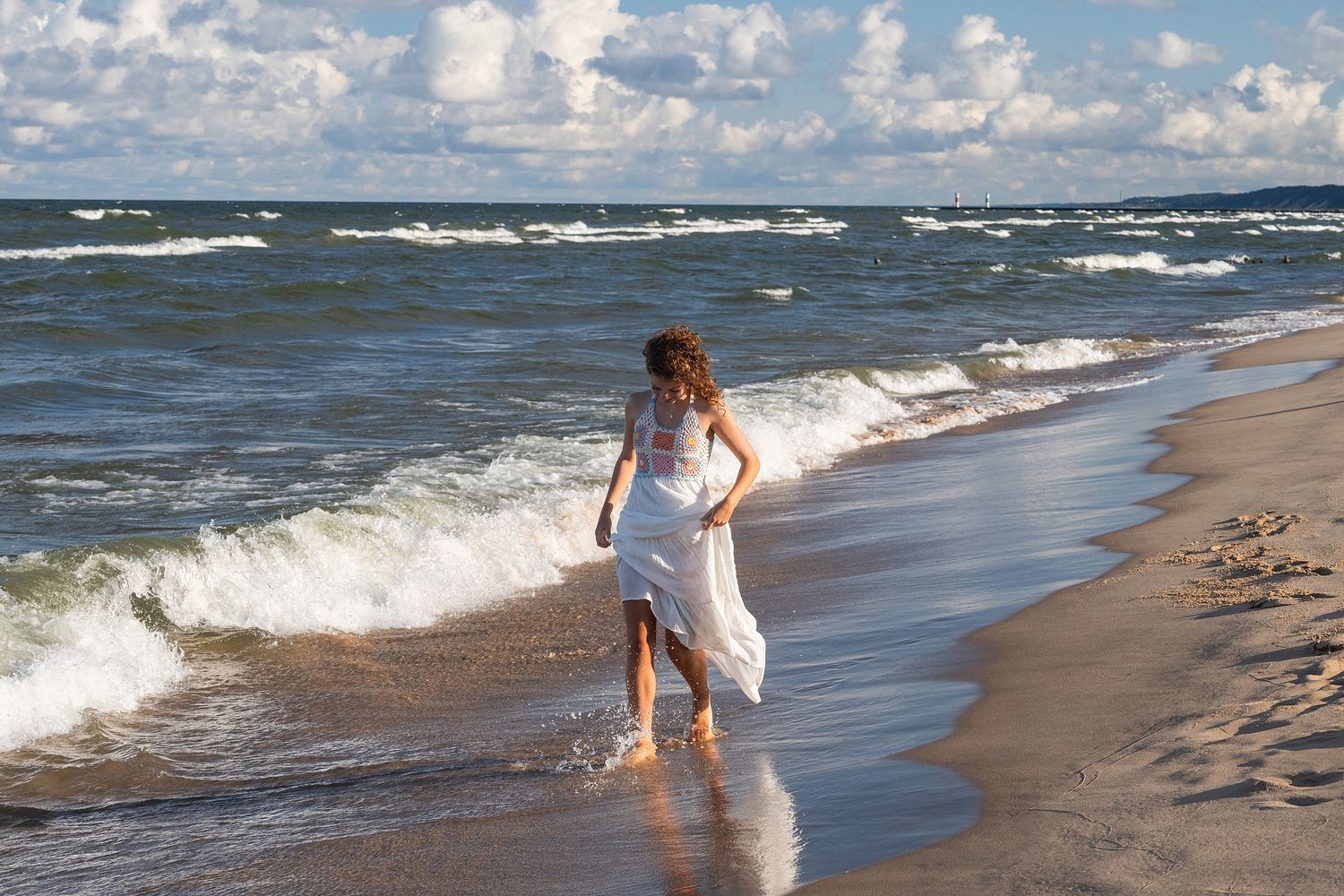 A figure in a white dress walks along a sandy beach with crashing waves and cloudy skies in the background.