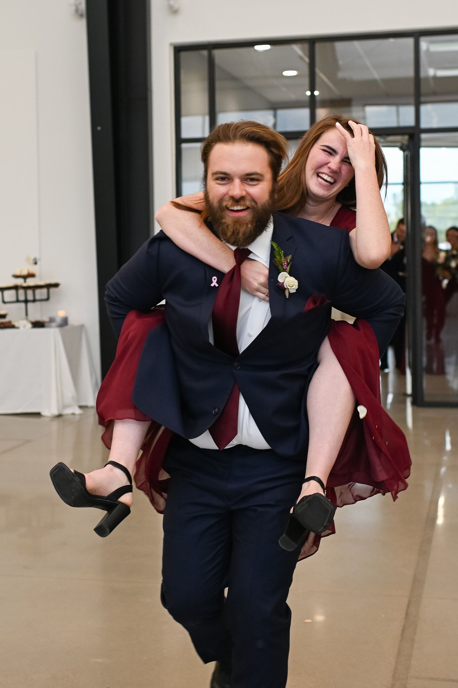 Groomsman giving bridesmaid a piggyback ride at wedding reception, both laughing joyfully in formal attire.