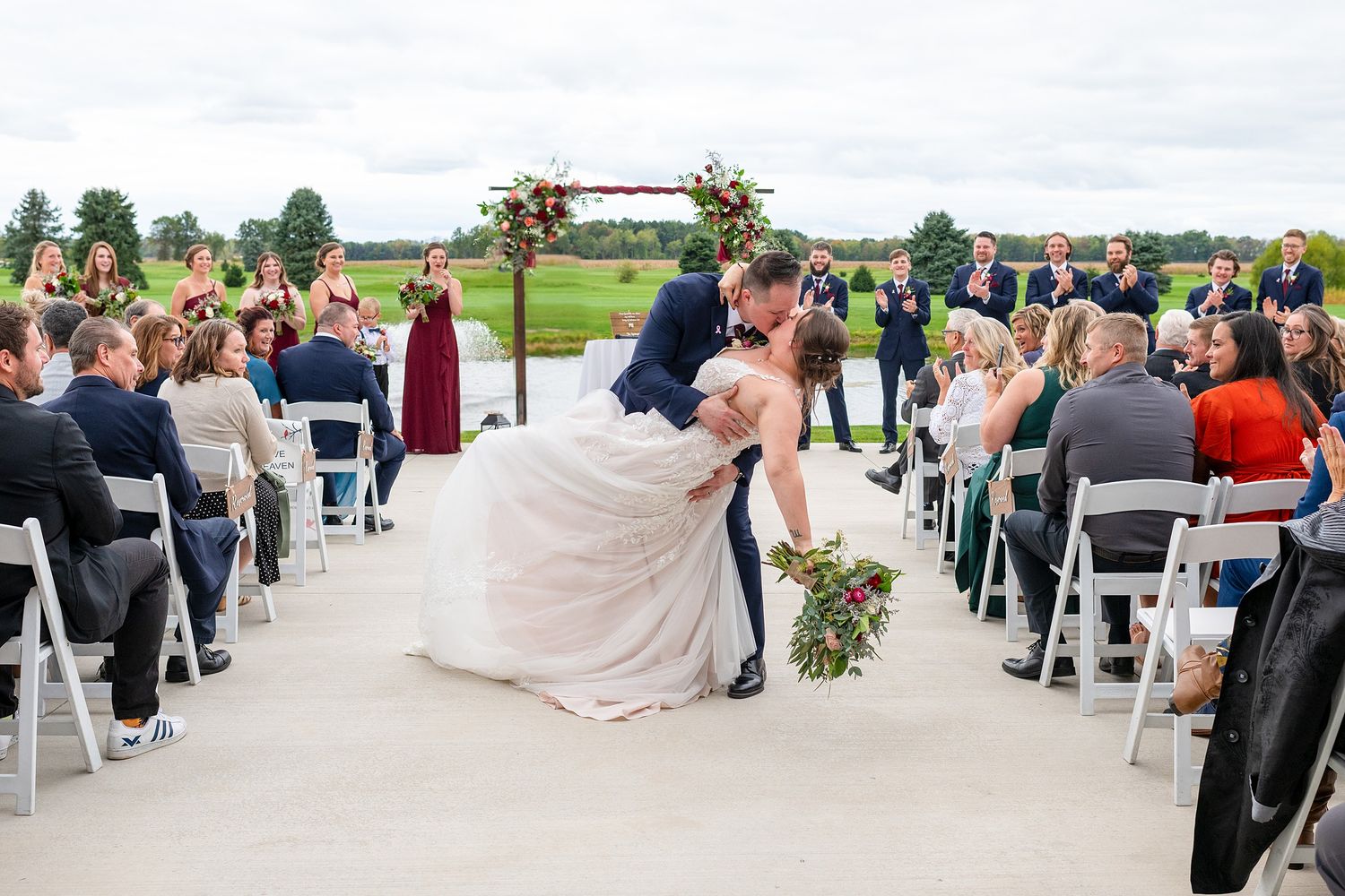 Groom dips bride for a romantic kiss at outdoor wedding ceremony aisle.