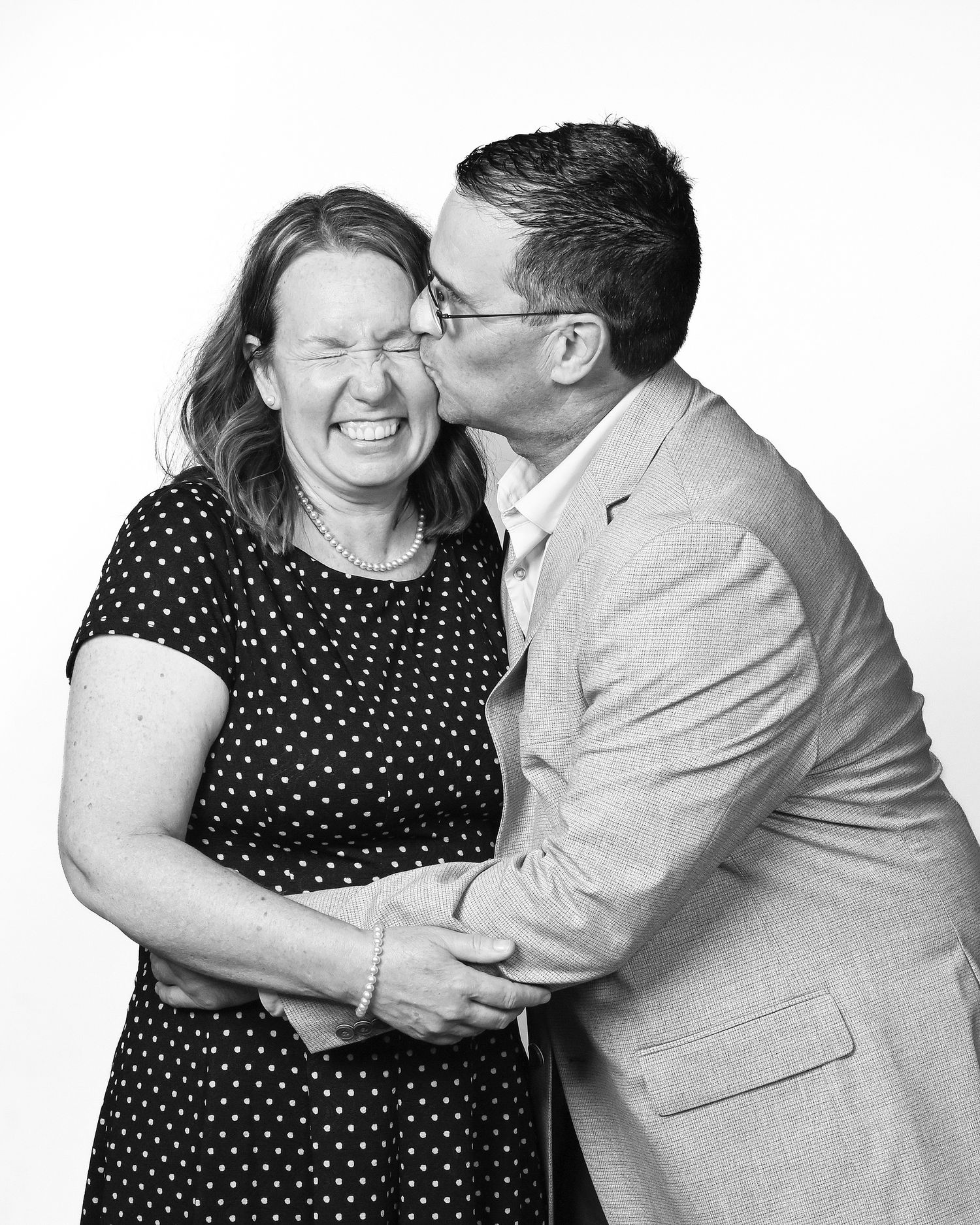 Black and white photo of a laughing woman being kissed on the cheek by a smiling man in a suit jacket.