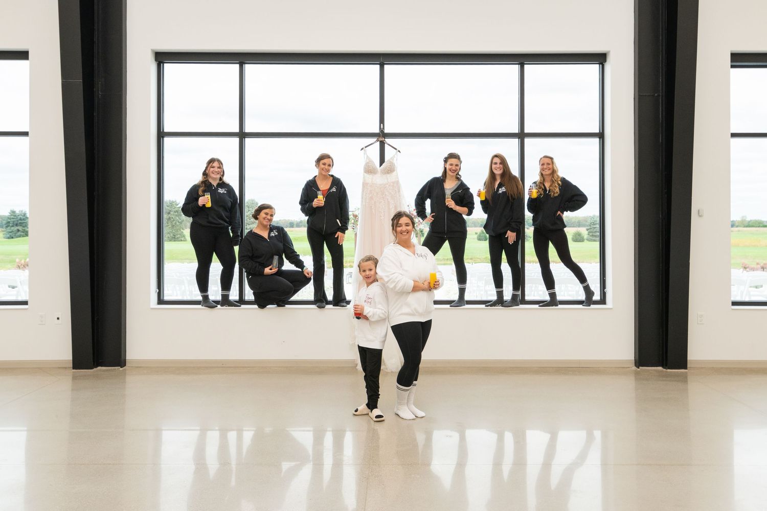 Bridal team in black posing with a wedding dress hanging in a bright studio with large windows.