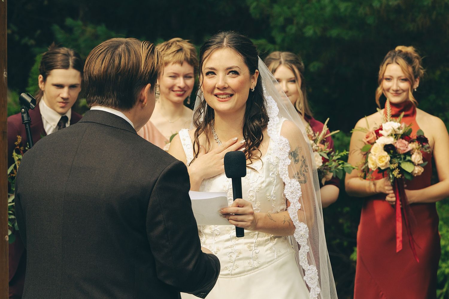 Smiling bride in a lace wedding gown and veil speaks into a microphone while holding vows, as bridesmaids with bouquets look on outdoors.