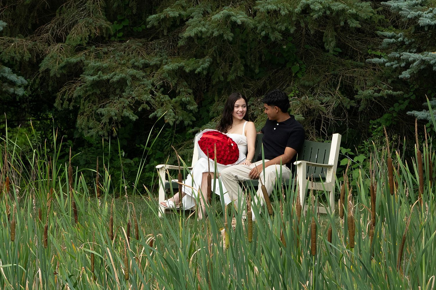 Couple sitting in white chairs surrounded by tall grass and evergreen trees in a lush garden setting.