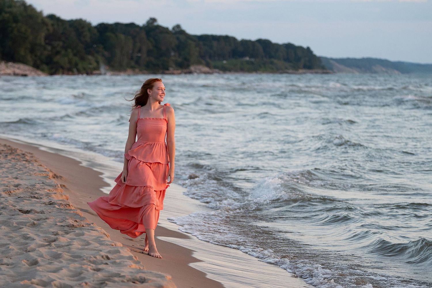 Woman in flowing coral dress walking barefoot along a sandy beach with waves and trees in background.