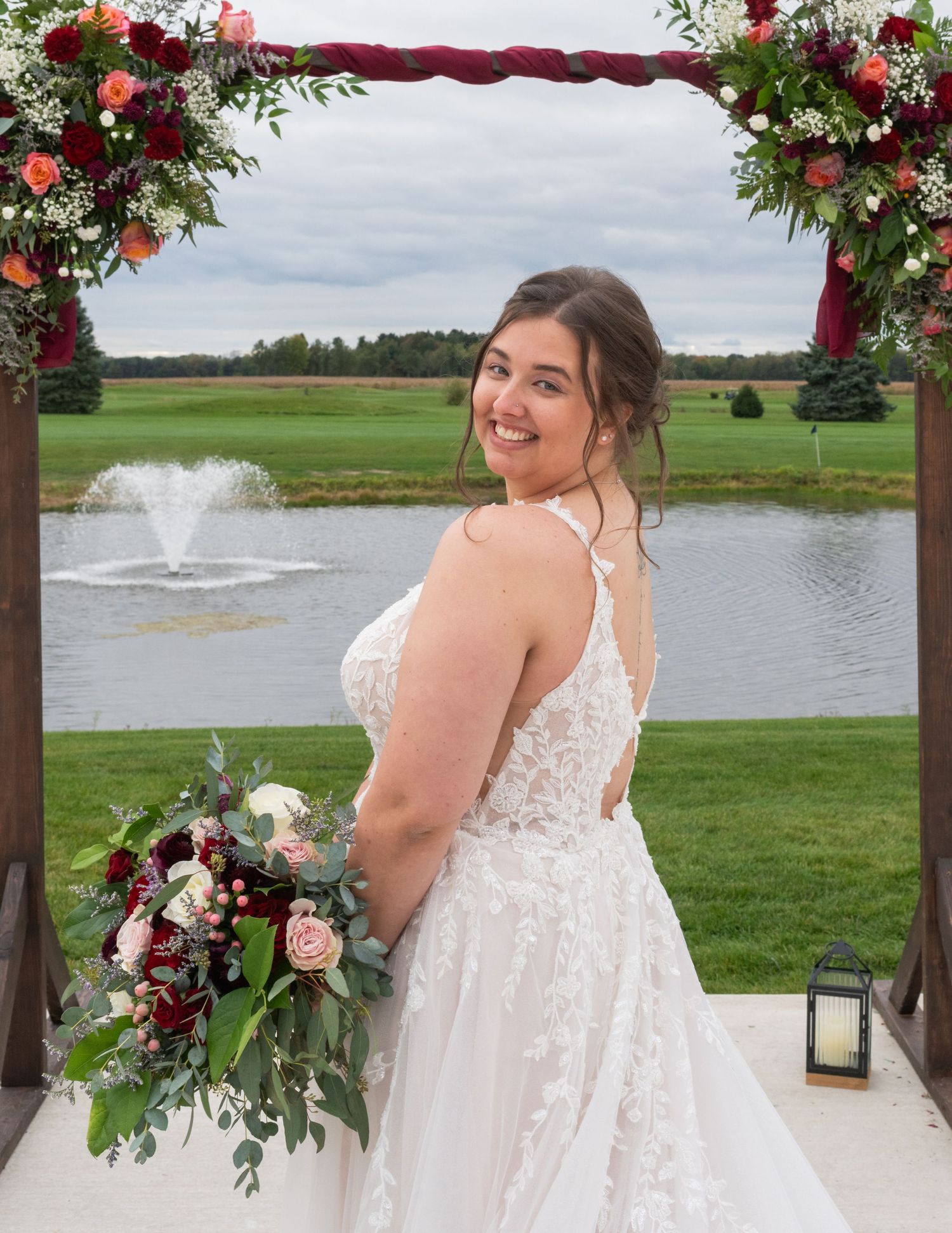 Smiling bride in lace wedding gown holding bouquet under floral arch with pond and fountain backdrop.