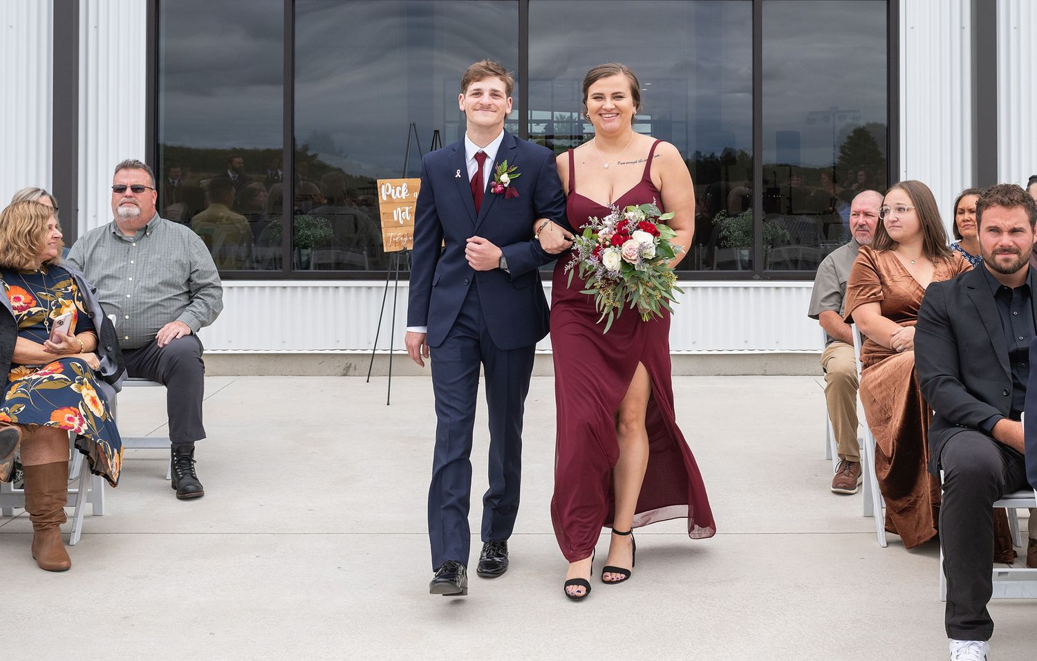 Groomsman escorts bridesmaid in burgundy gown down outdoor wedding aisle while guests watch.