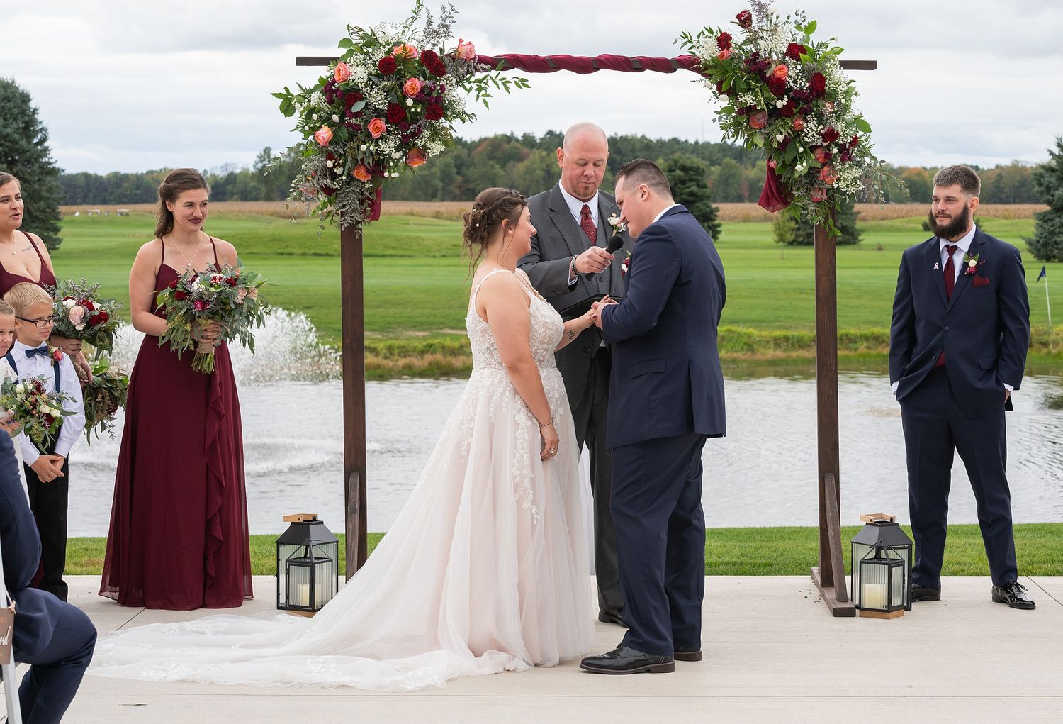 Groom placing ring on brides finger during outdoor wedding ceremony beside scenic pond and arch.
