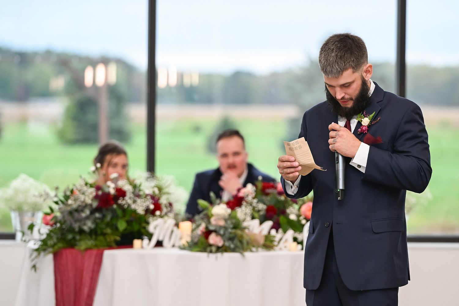 Groomsman in navy suit delivering heartfelt speech at wedding reception while reading from notes.