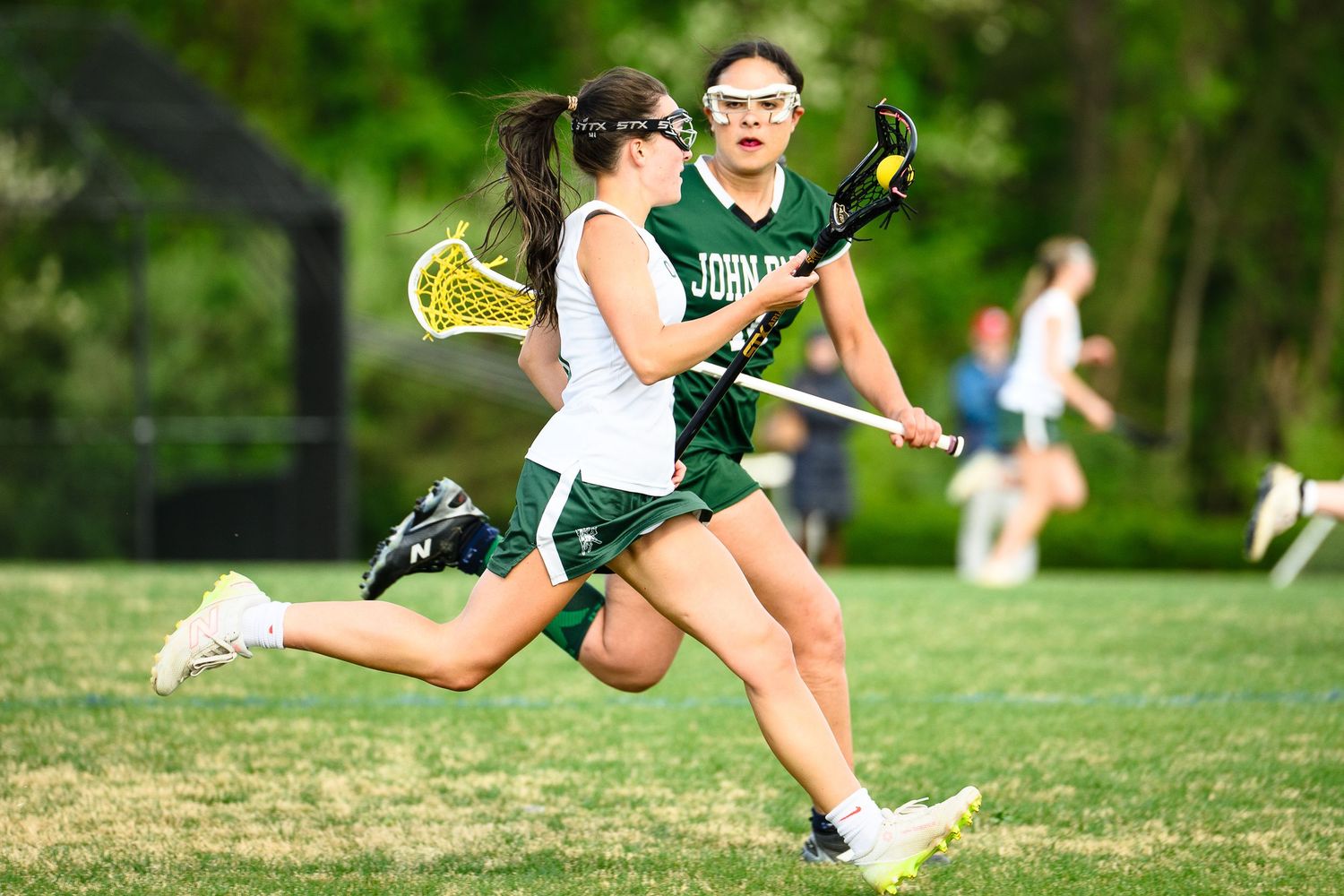 Female lacrosse player in white uniform sprints past defender in green during an outdoor game.