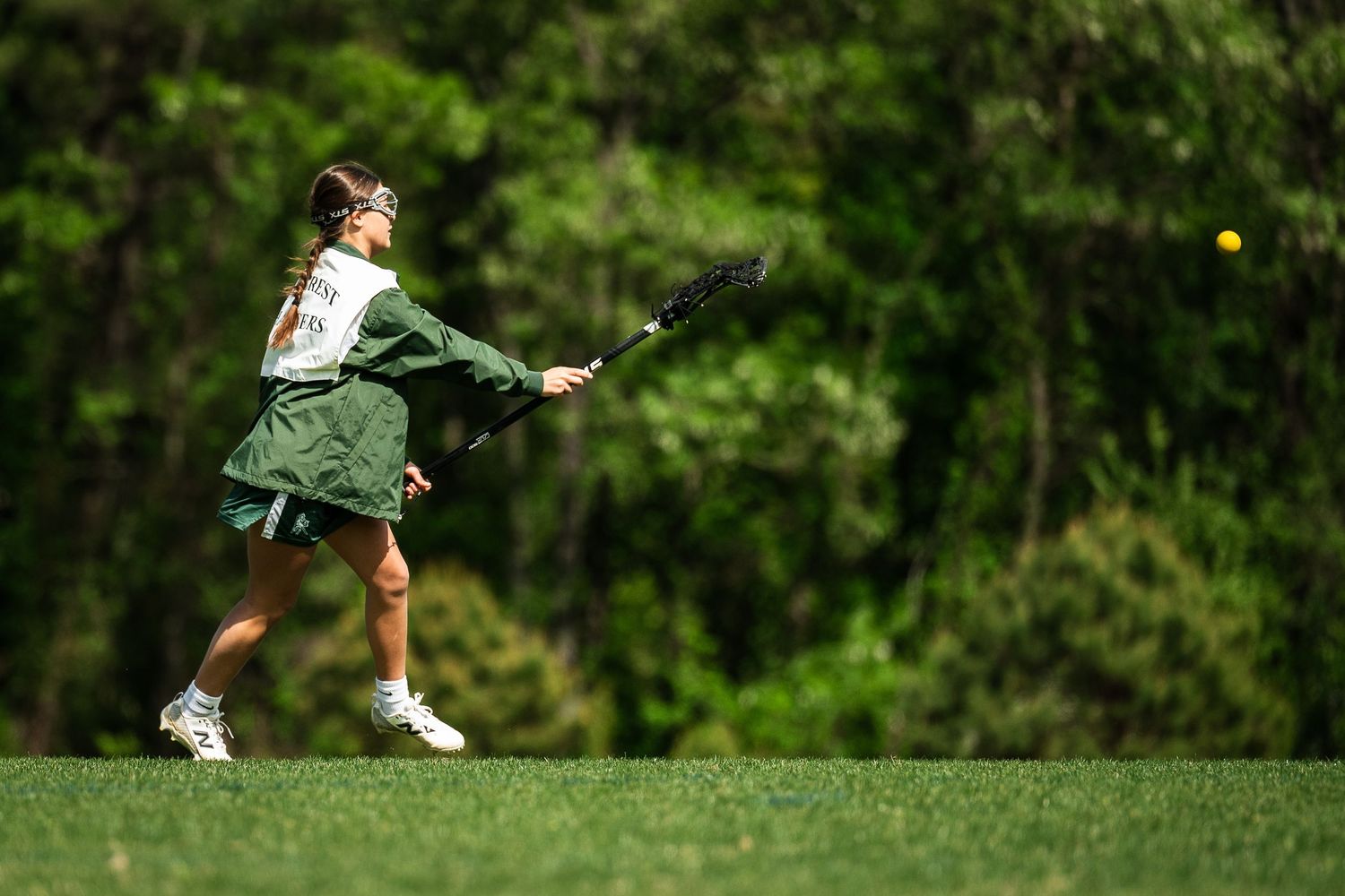Female lacrosse player in green uniform swinging stick on grass field with trees in background.