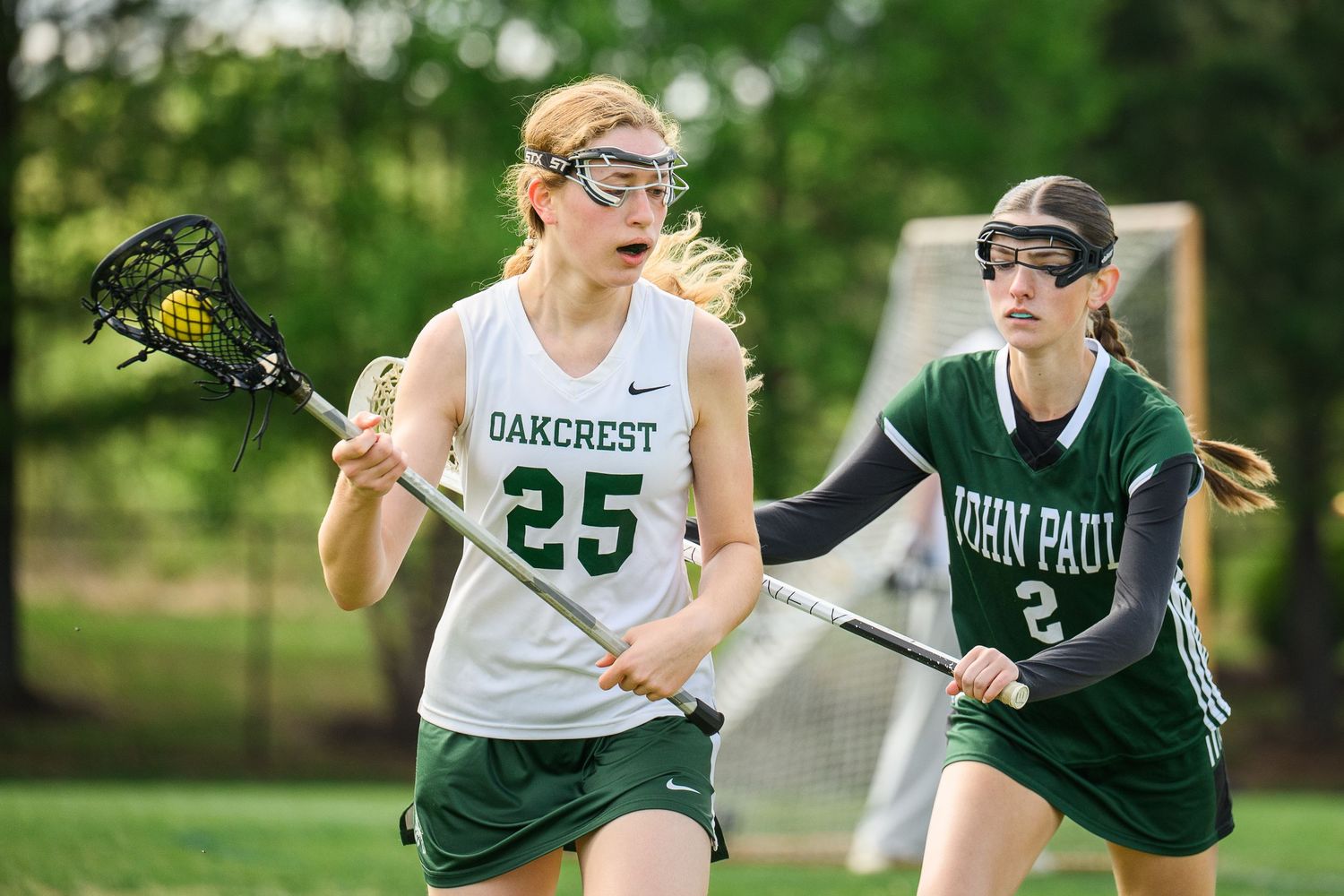 Two female lacrosse players compete, one in Oakcrest #25 jersey, the other in John Paul #2, on a green field.