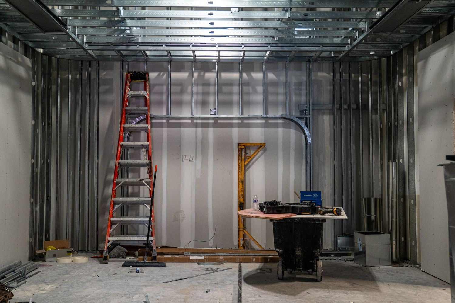 Construction site interior with metal framing, orange ladder, and temporary work table against unfinished walls.