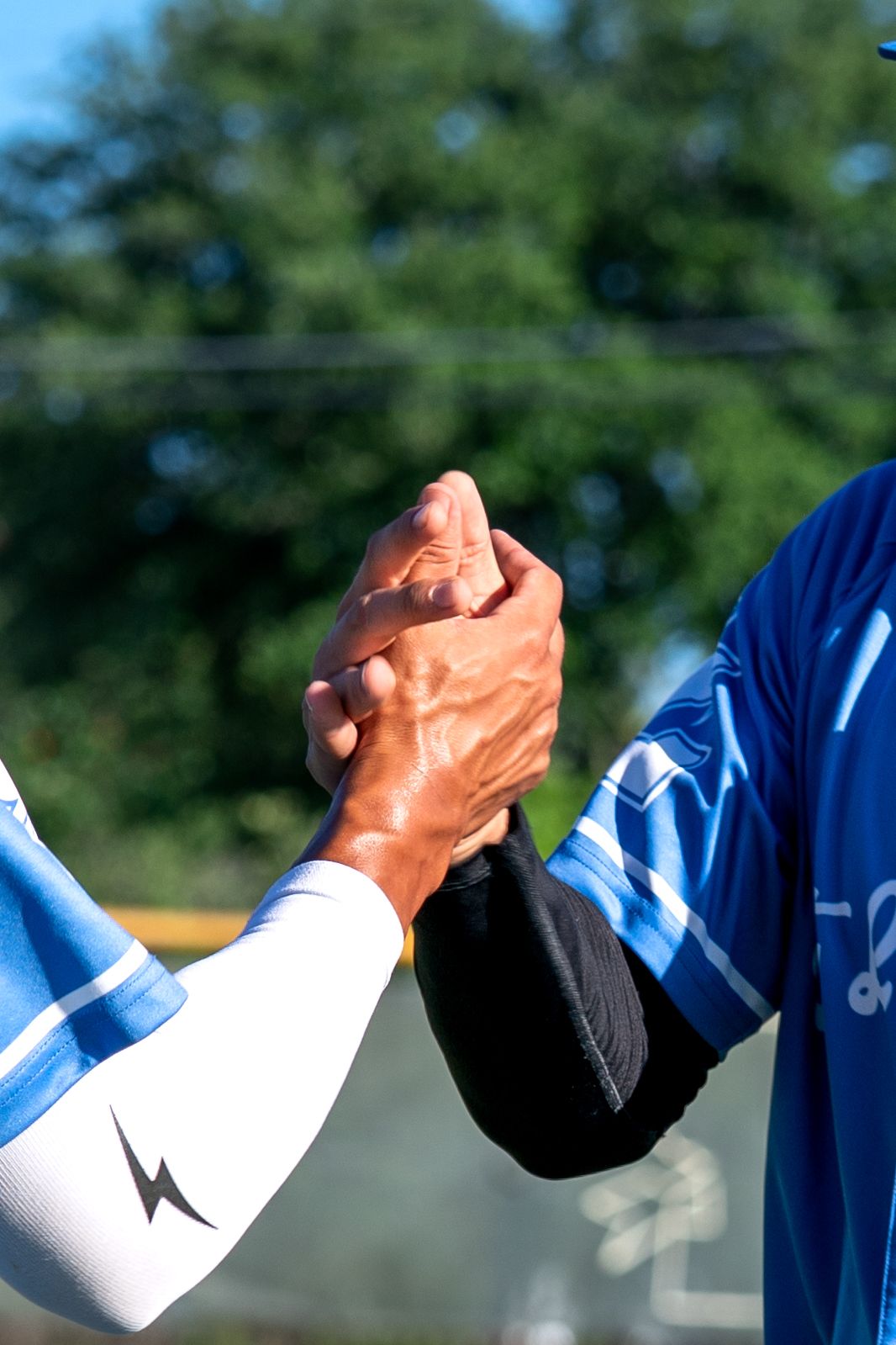 Two athletes in blue and white jerseys shake hands outdoors after a sporting event, with green trees in the background.
