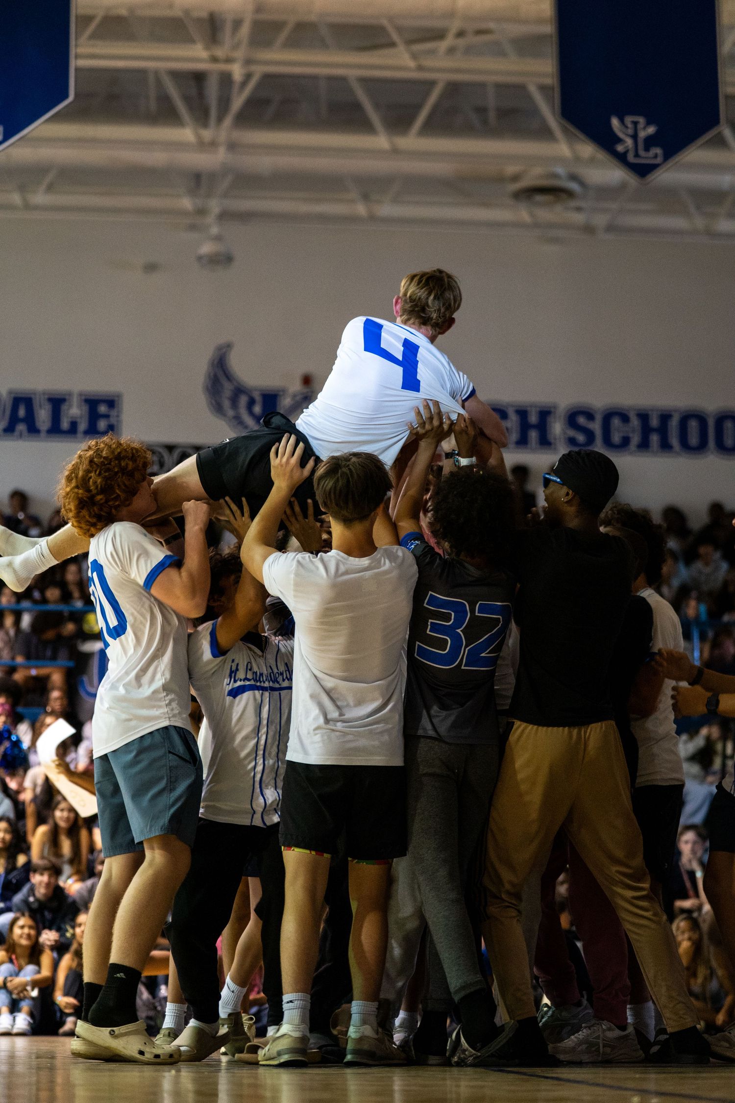 Students lift teammate wearing jersey number 4 during celebratory moment in high school gymnasium.