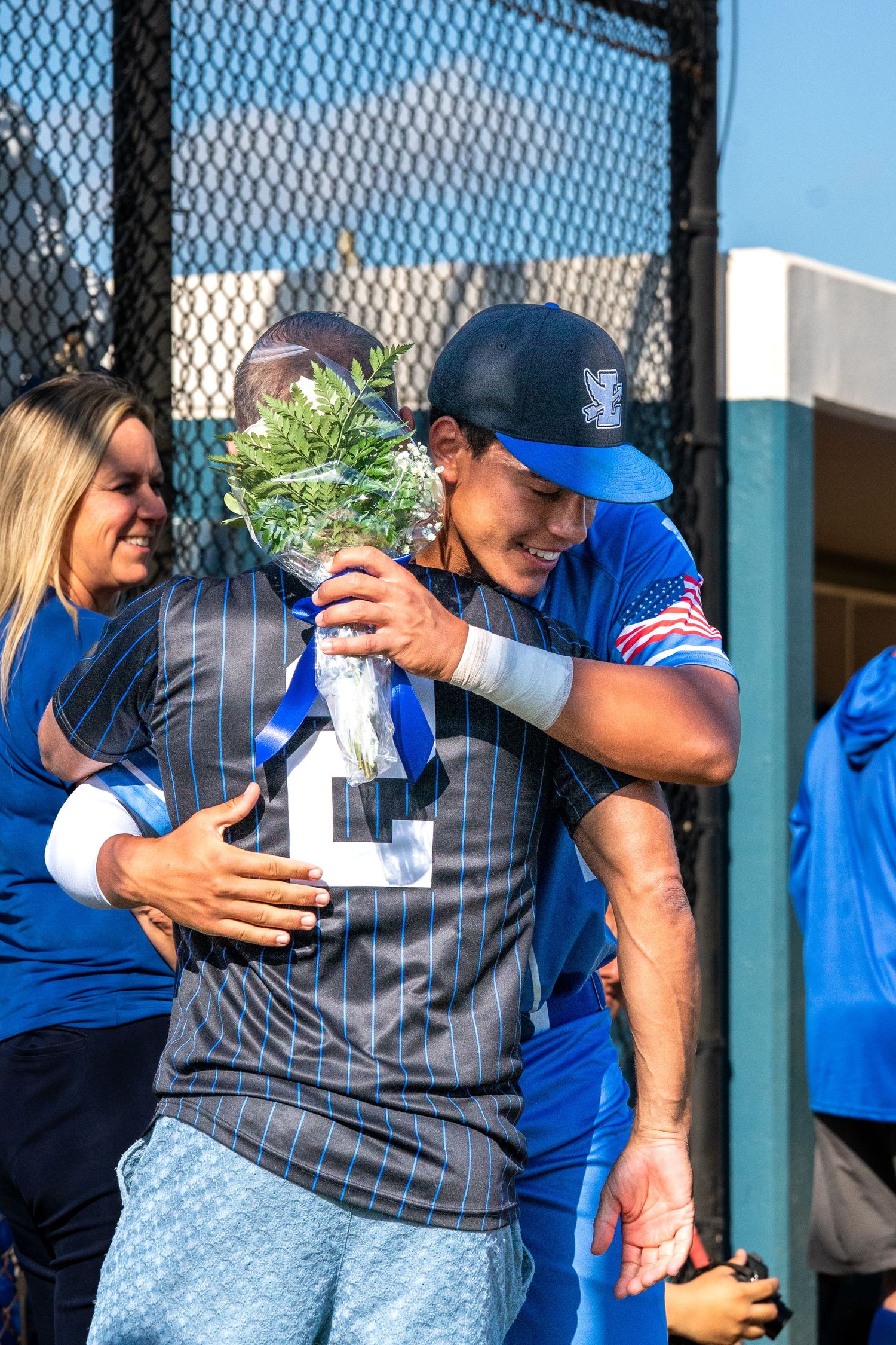 Baseball player in blue uniform hugging teammate holding flowers during senior day ceremony on field.