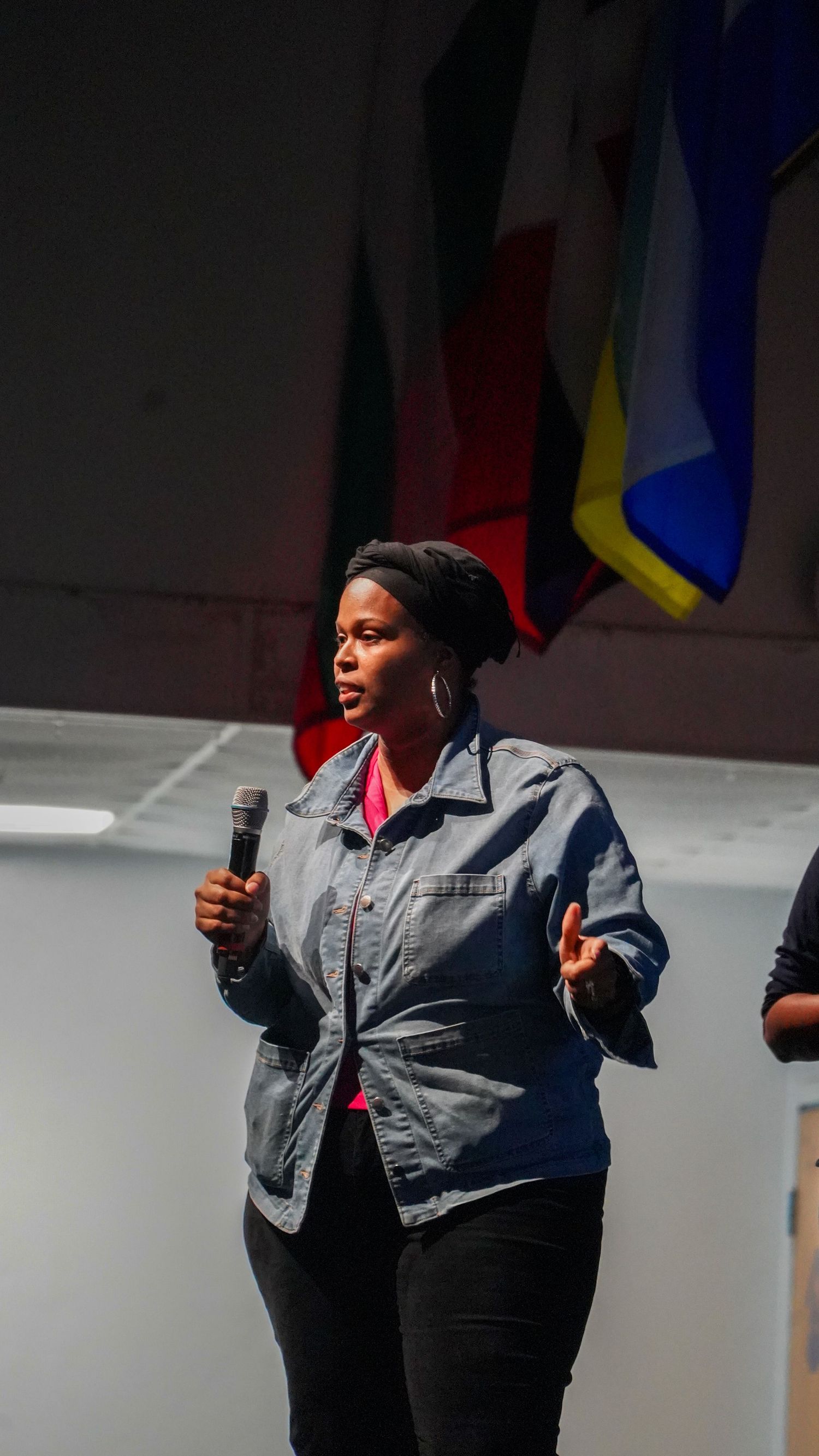 Speaker in a denim jacket gestures while presenting in front of colorful flags.
