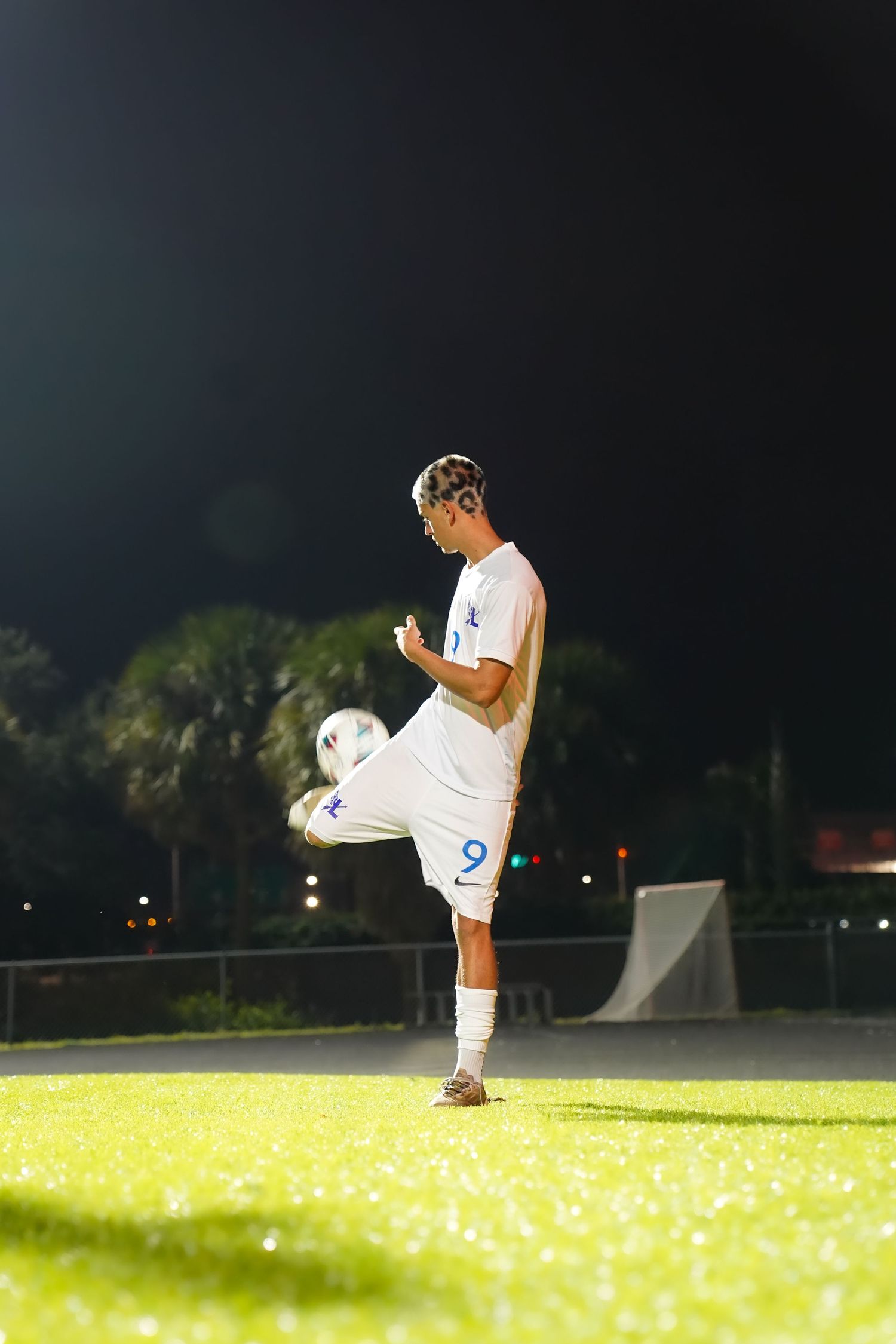 Soccer player practicing juggling skills on illuminated grass field at dusk.